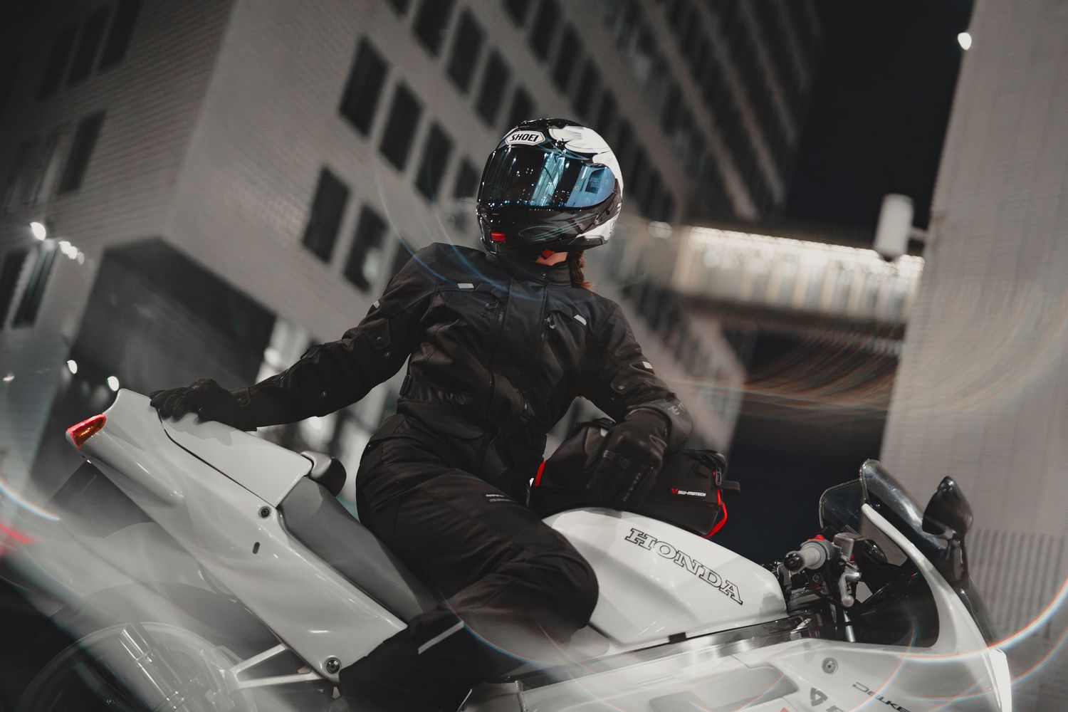 A motorcyclist in a black jacket and helmet rides a white sport motorcycle through an urban setting at night.