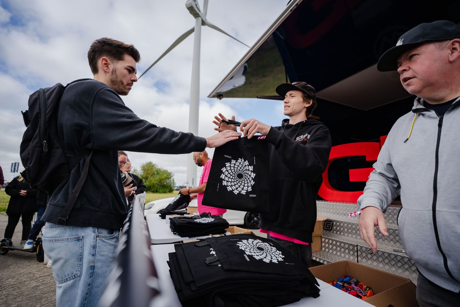 Vendor selling branded black t-shirts with white logo design at outdoor merchandise booth near wind turbine.