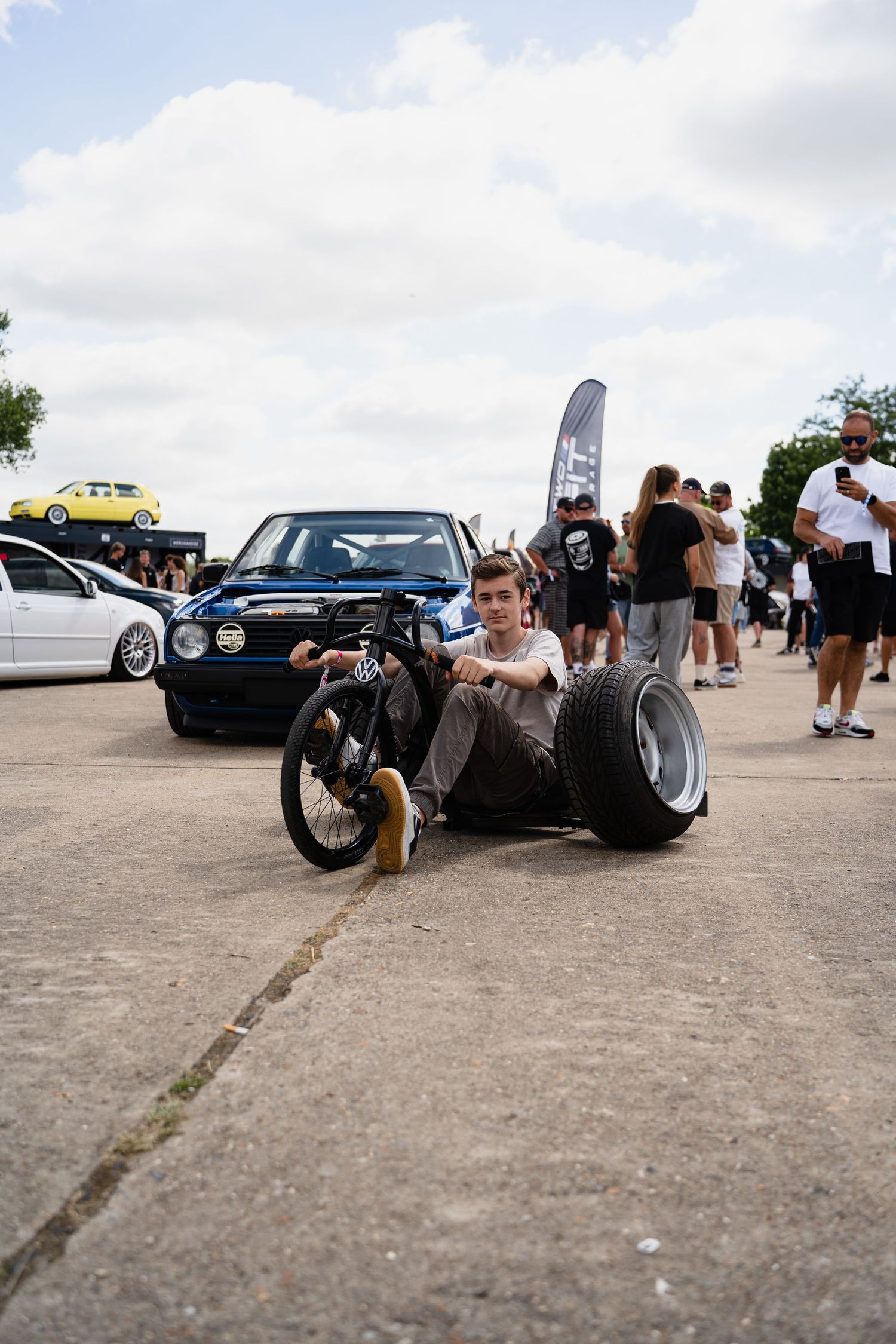 Boy sits on ground with bicycle wheels at outdoor car show, with classic VW Golf and spectators in background.