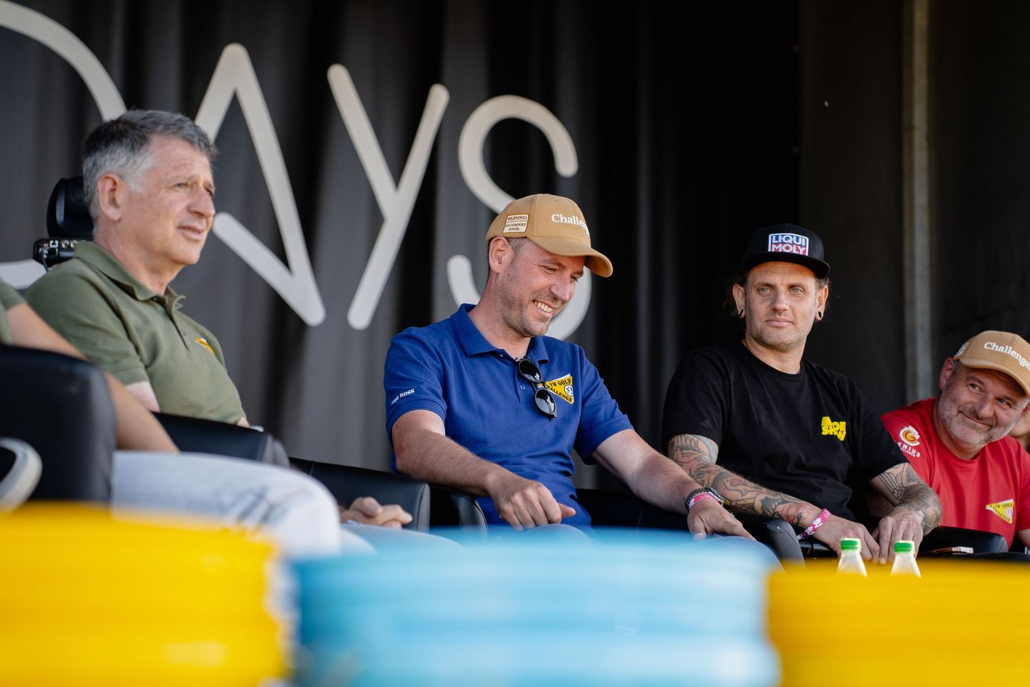Four men sitting together at an outdoor event booth, smiling and chatting in casual attire.