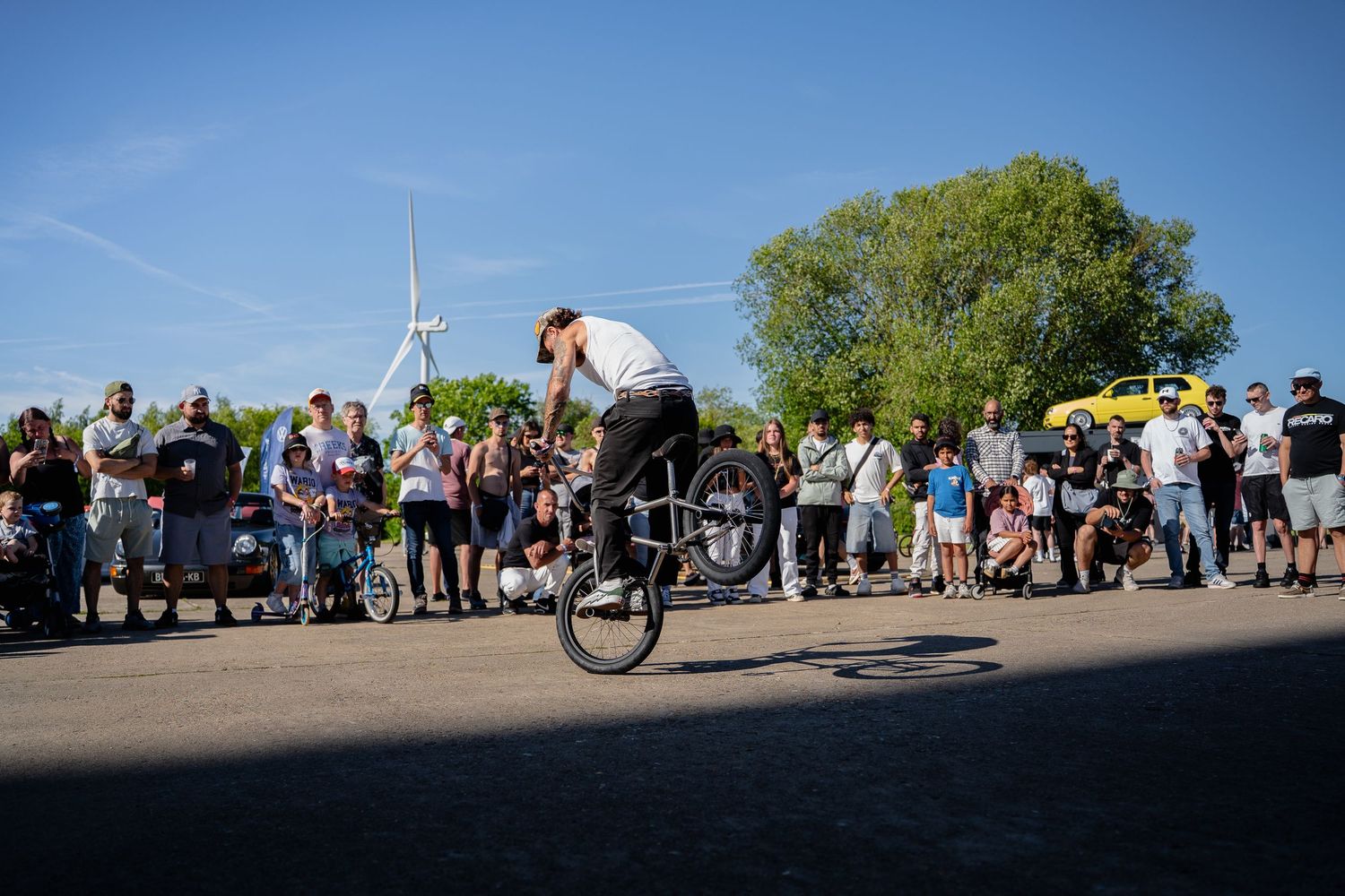 BMX rider performs a wheelie trick for a crowd of spectators outdoors near trees and a wind turbine.