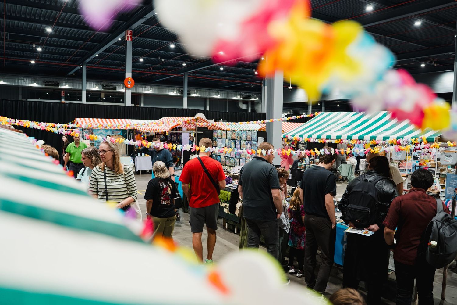 Busy indoor market with colorful balloon decorations, striped vendor tents, and crowds of shoppers browsing merchandise.