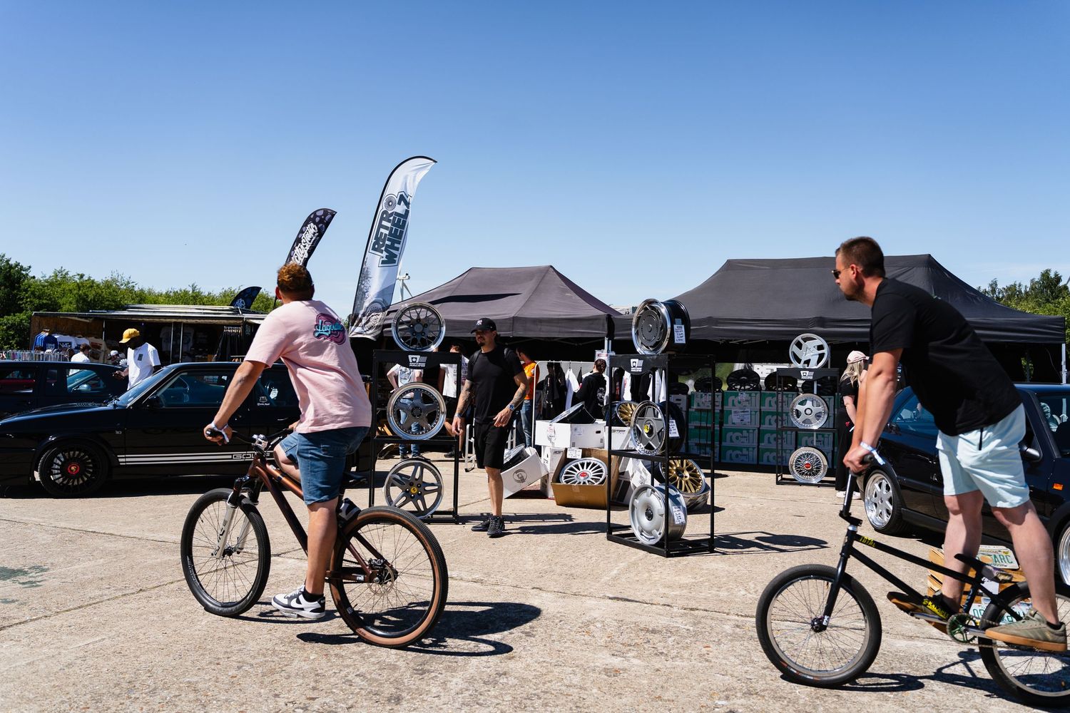 Two men riding BMX bikes at an outdoor cycling event with black tents and display stands on a sunny day.