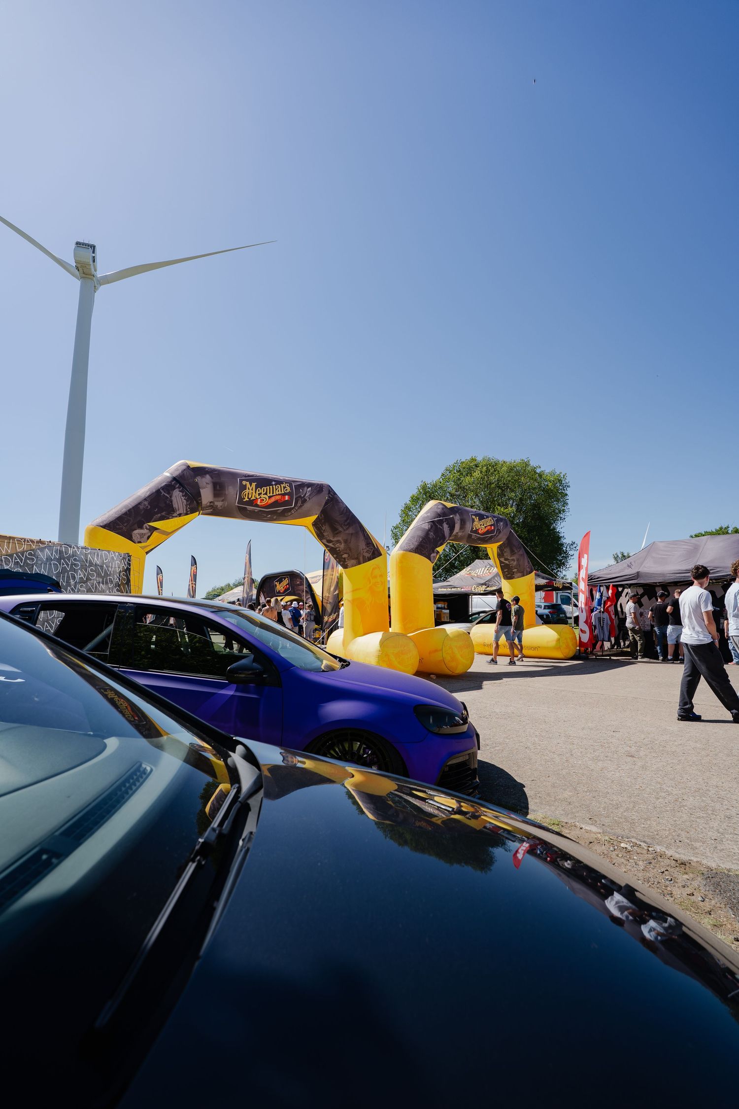 Meguiar's inflatable arch at an outdoor car show with a blue VW, wind turbine, and crowds under clear blue sky.