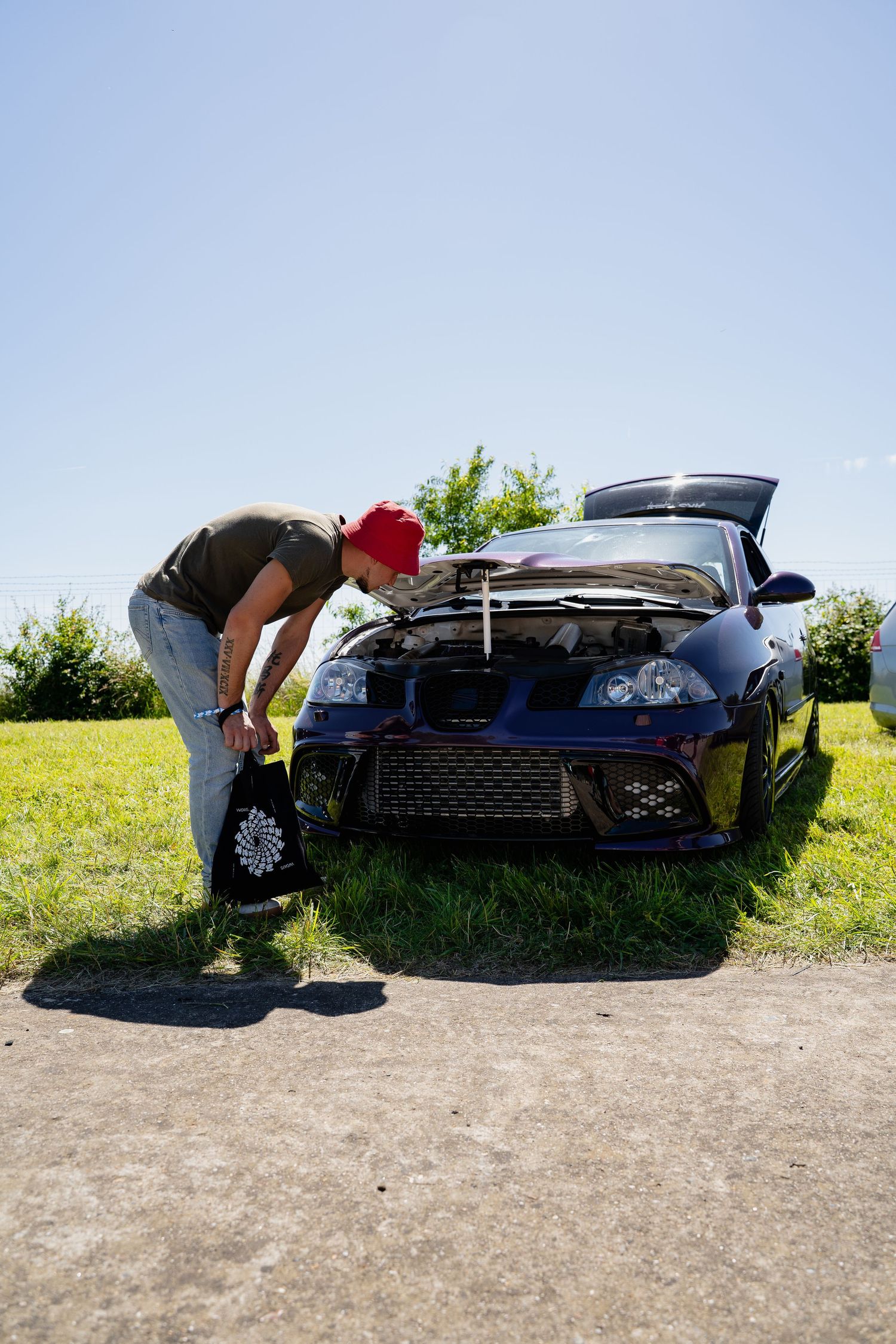 Man in red cap inspecting open hood of dark blue sports car on sunny day outdoors.