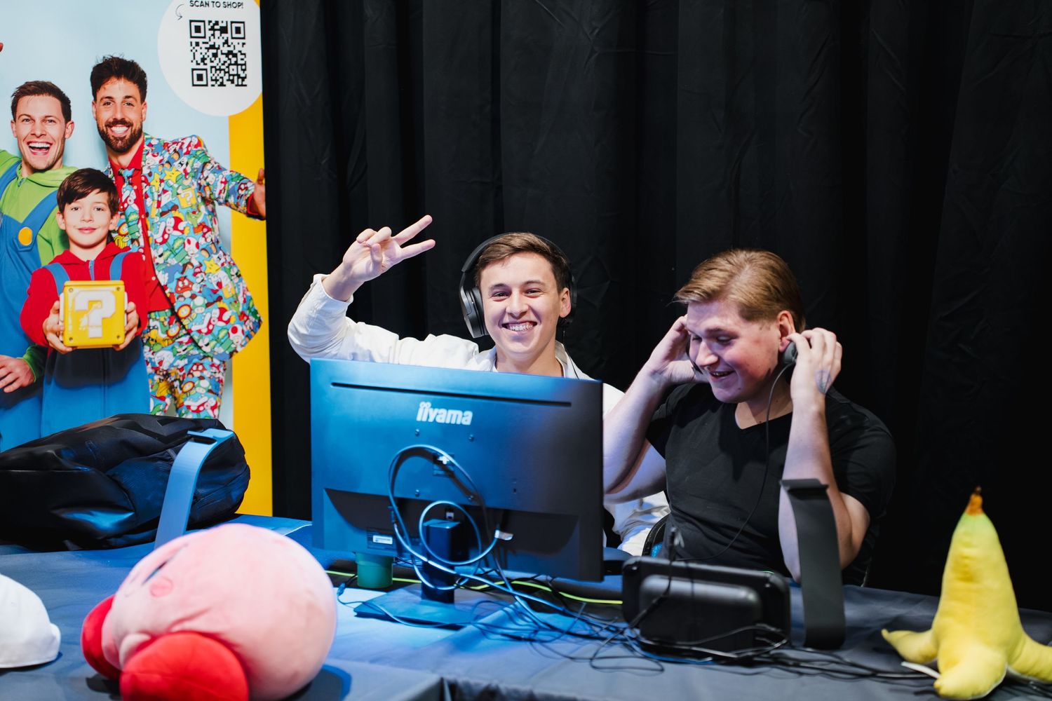 Two gamers smile at a PC setup with Kirby plush and banana toy at a gaming event.