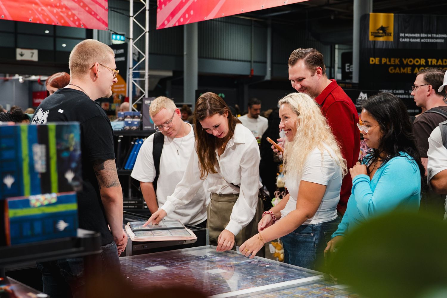 Group of people gathered around a gaming table at an indoor expo or gaming convention event.