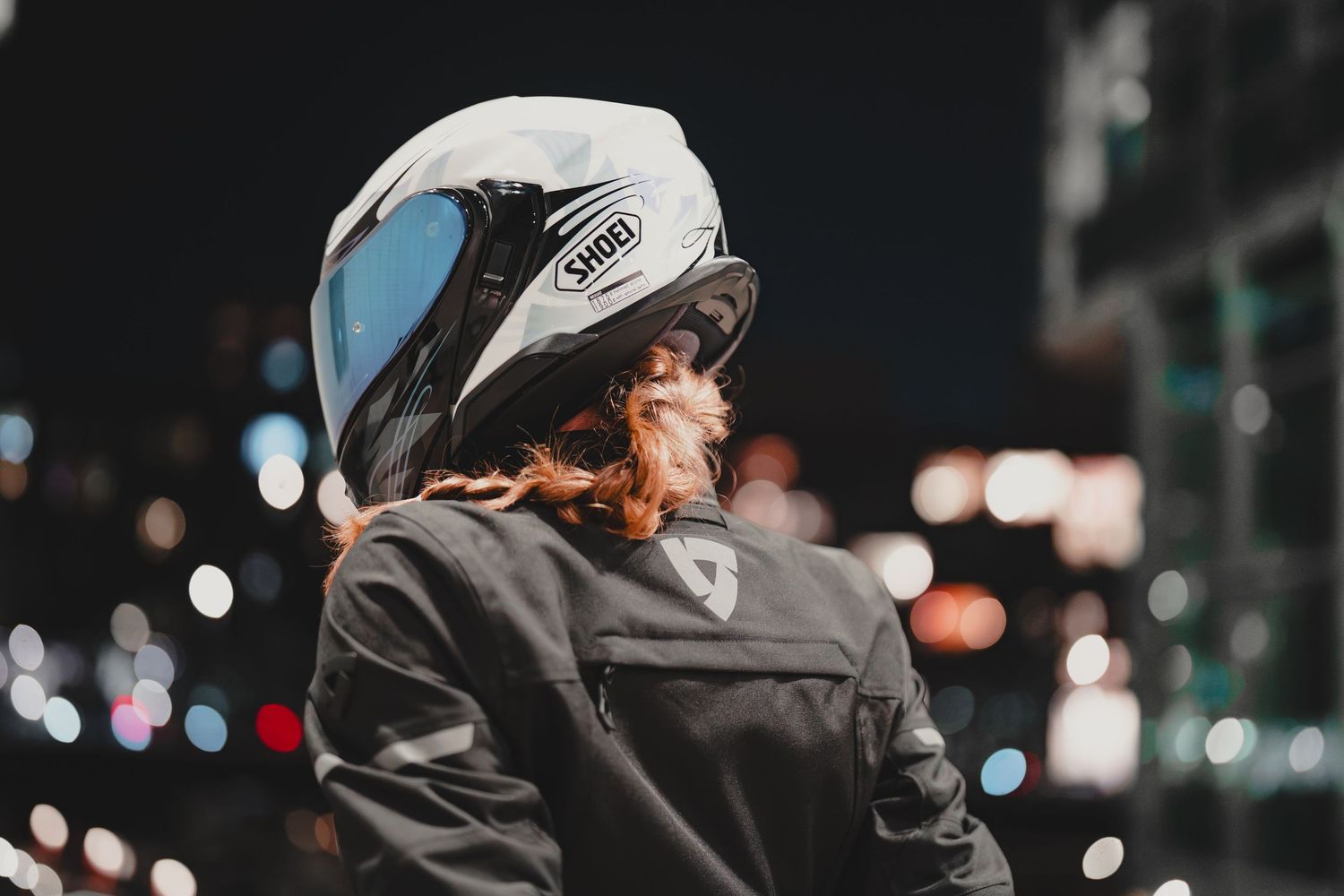 Person wearing a white Shoei motorcycle helmet and black riding jacket at night with bokeh city lights in background.