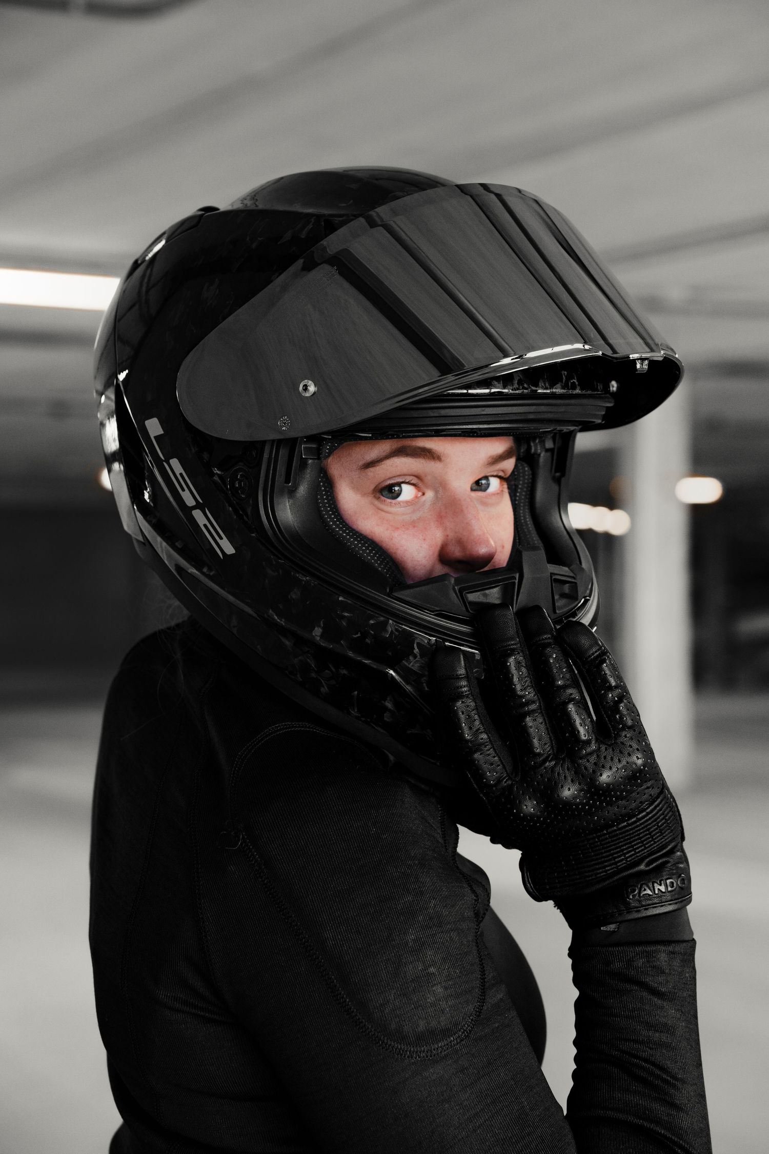 Person wearing black motorcycle helmet and gloves in a parking garage, looking sideways with visor up.