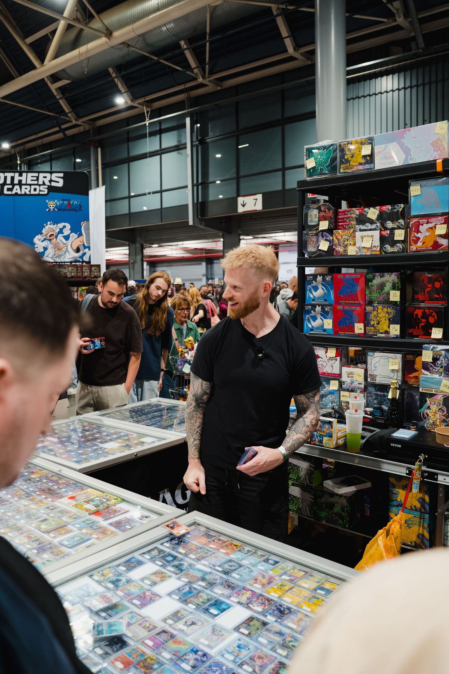 Man with tattoos browsing trading cards at a busy convention vendor booth filled with collectibles.