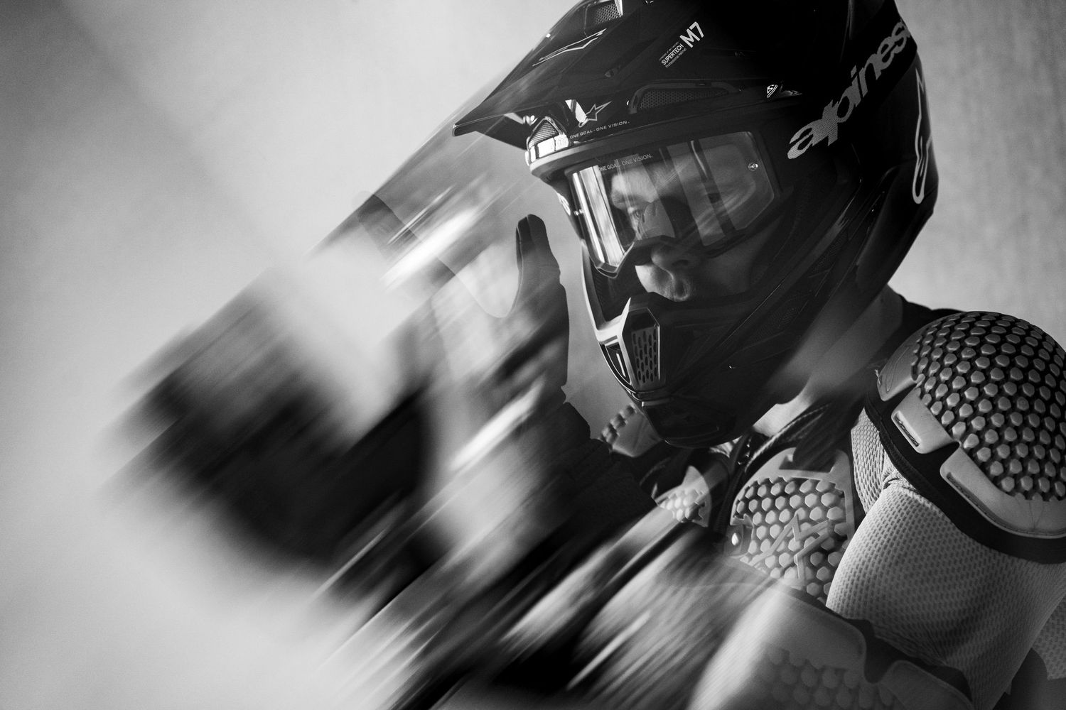Black and white photo of a motocross rider wearing a helmet and protective gear, with motion blur effect.
