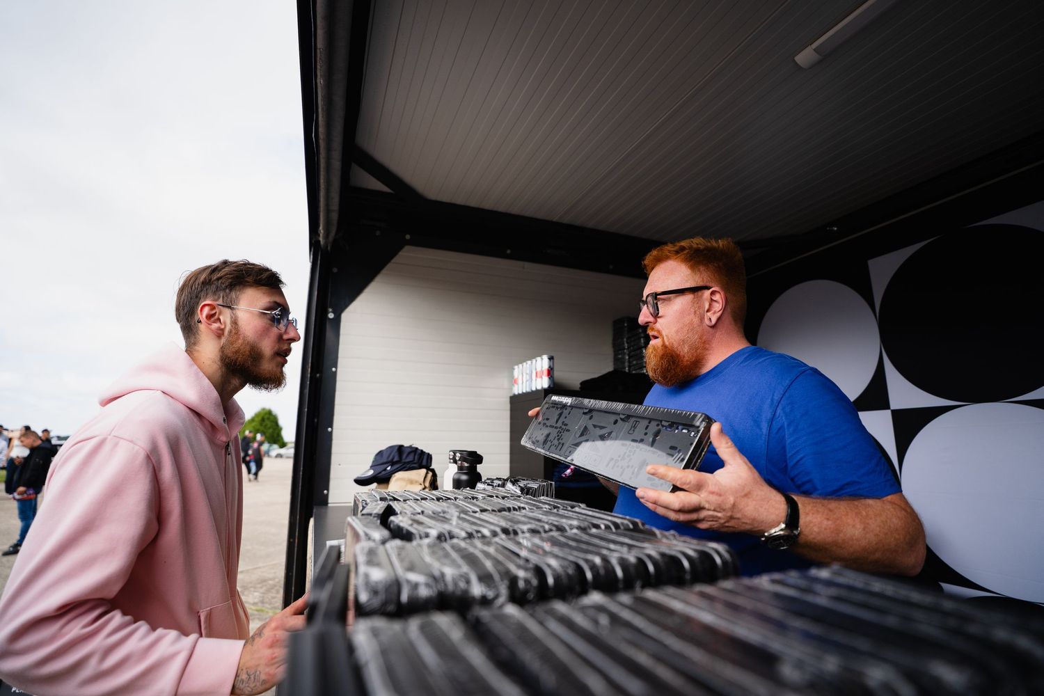 Two men talking near open truck trunk with vinyl records at outdoor event.