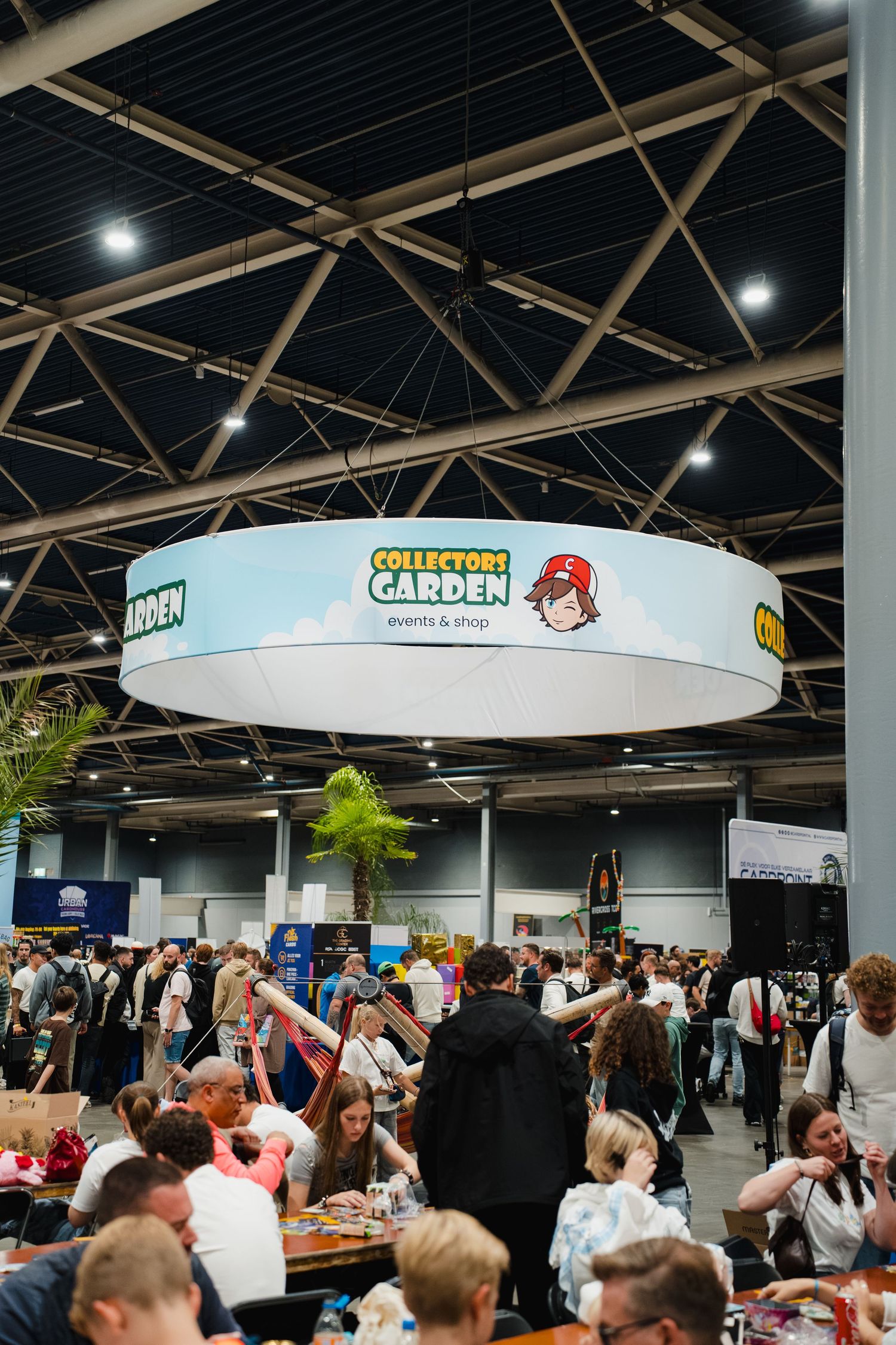 Crowded Collectors Garden events and shop booth inside a large convention hall with a circular hanging sign.