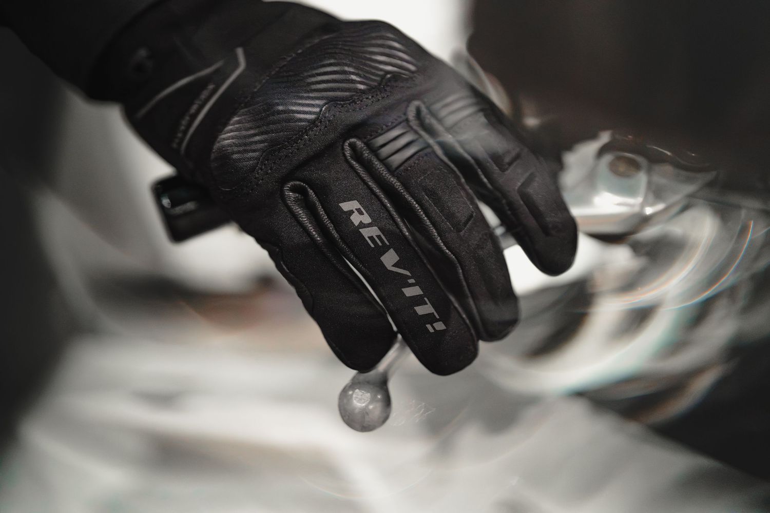 Black REV'IT motorcycle glove holding a small metal ball, showcasing grip and protective design.