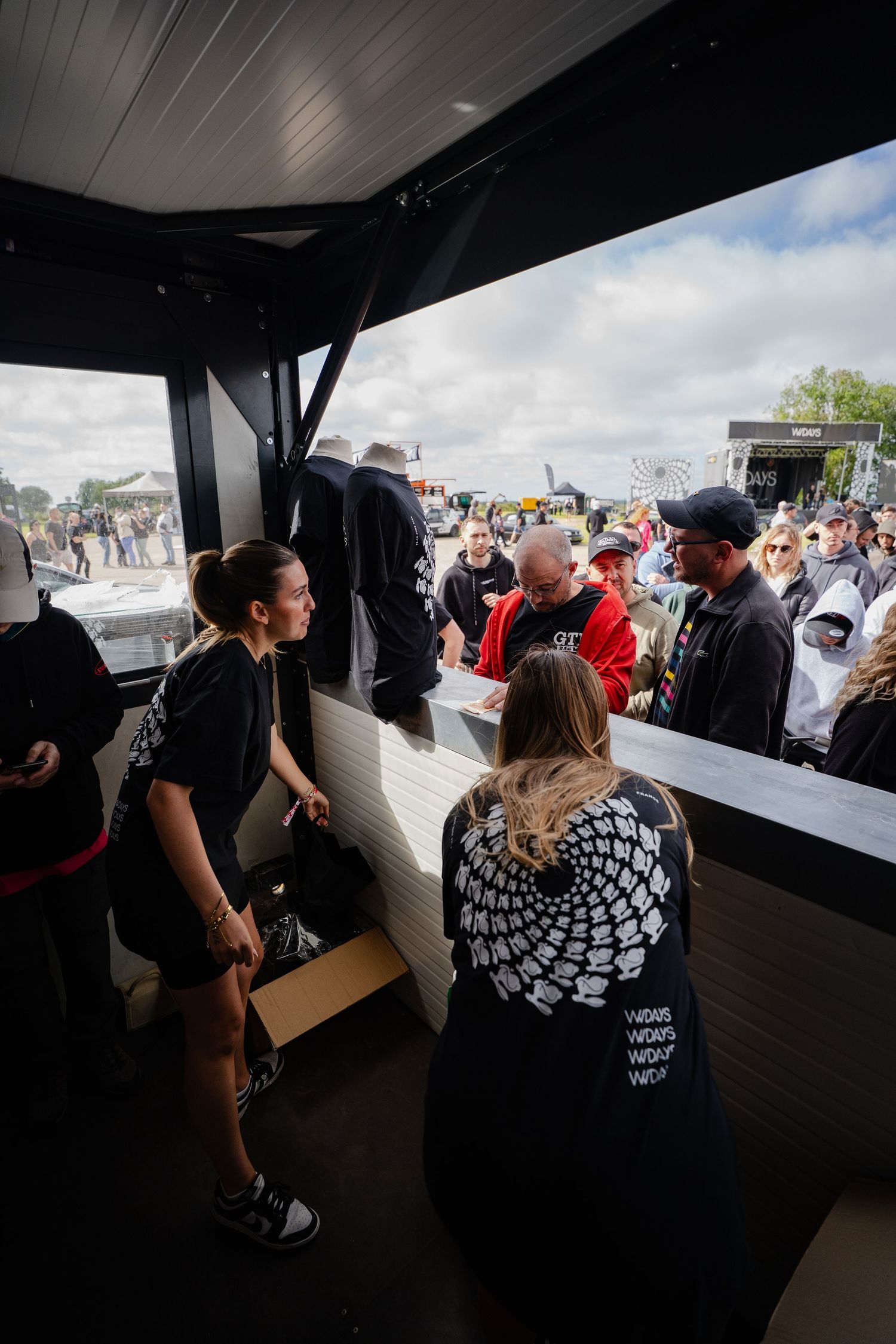 People gathered at an outdoor festival vendor booth, with staff serving customers under a dark canopy on a cloudy day.