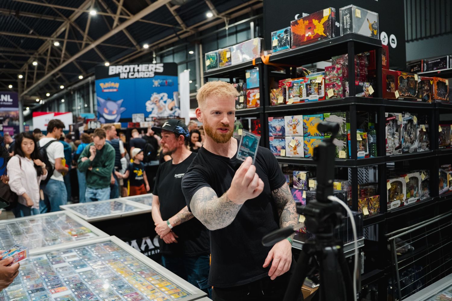 Tattooed man at a gaming convention booth browsing collectible cards with crowded vendor displays behind him.