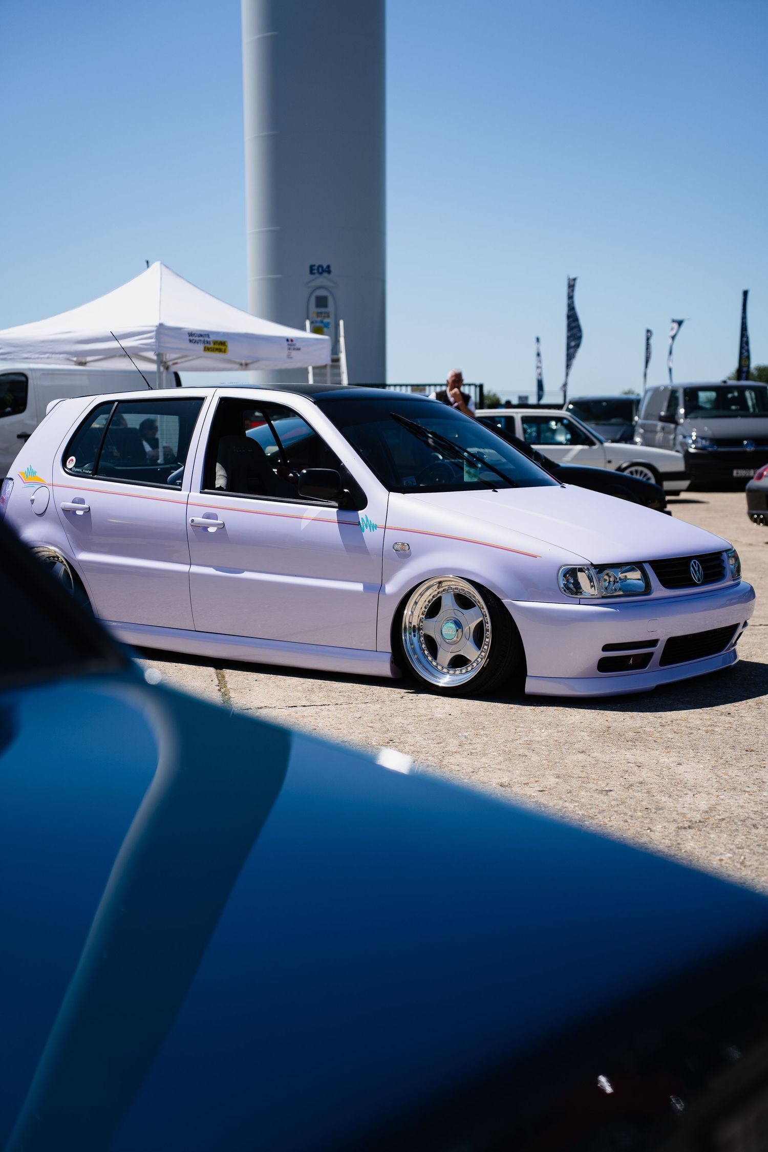 A lowered white Volkswagen Polo at a car show, featuring custom wheels and a slammed stance on a sunny day.
