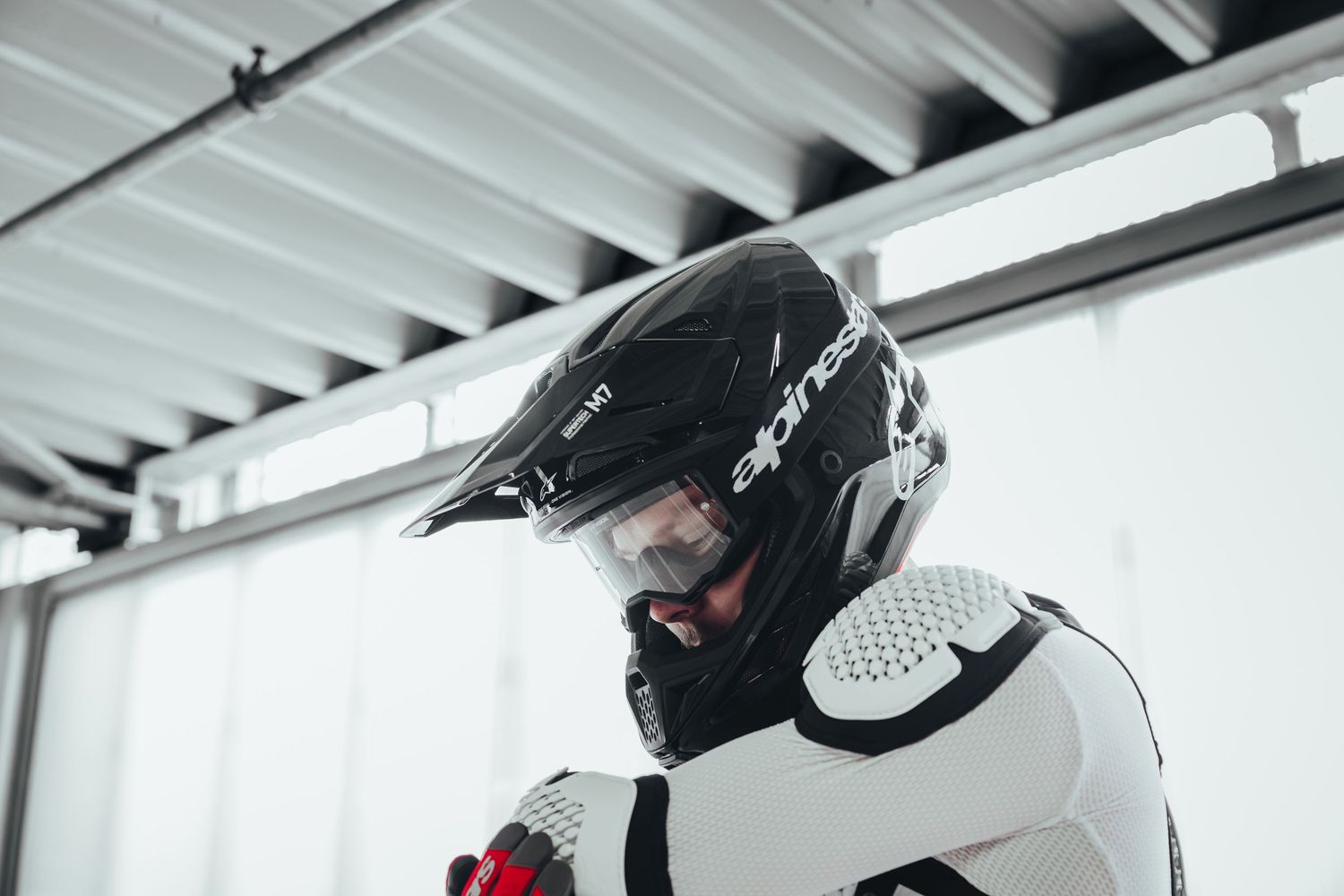 Motocross rider wearing a black Alpinestars helmet and white protective gear standing indoors.