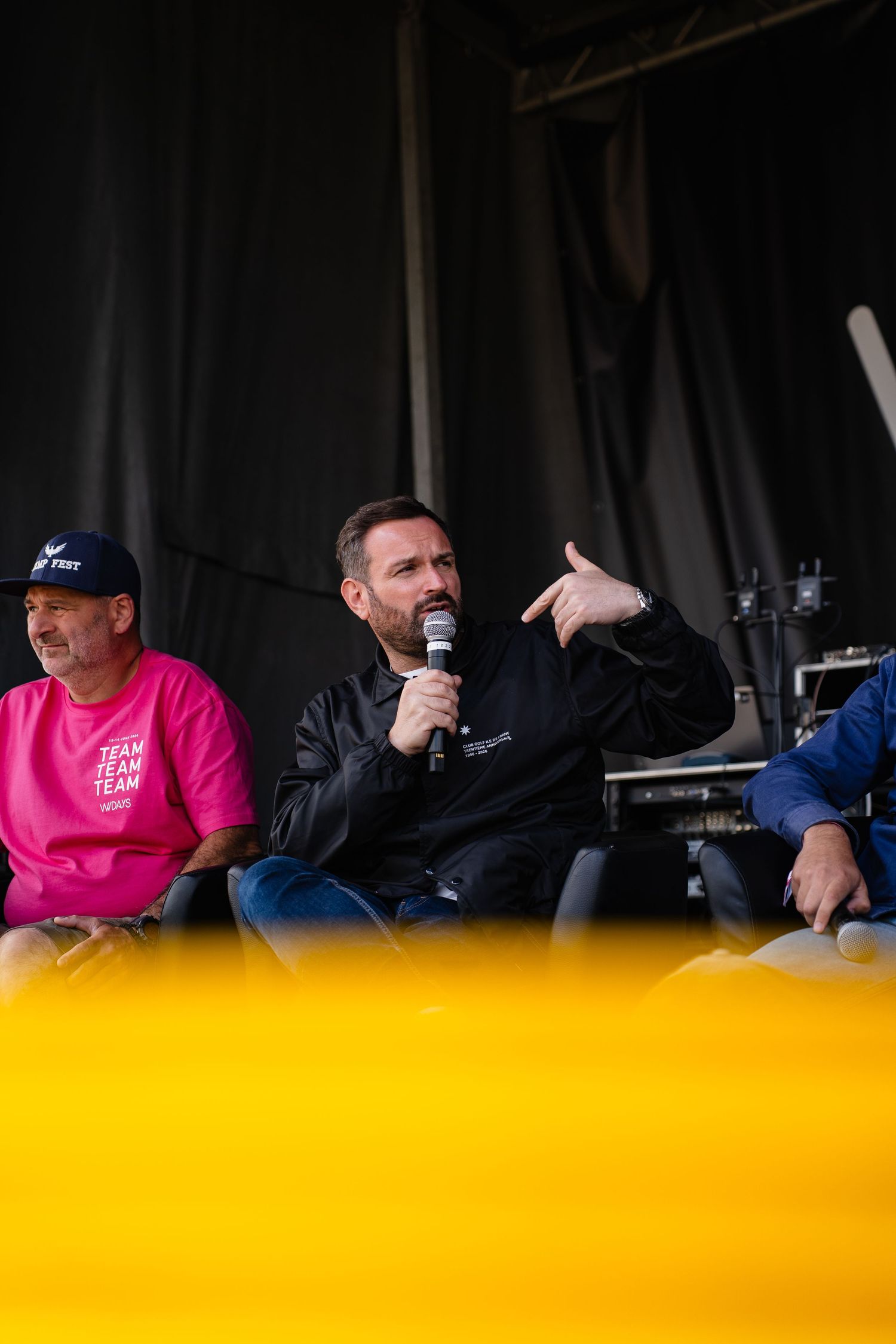 Three men sit on stage at an outdoor event, center man speaking into a microphone in a black jacket.