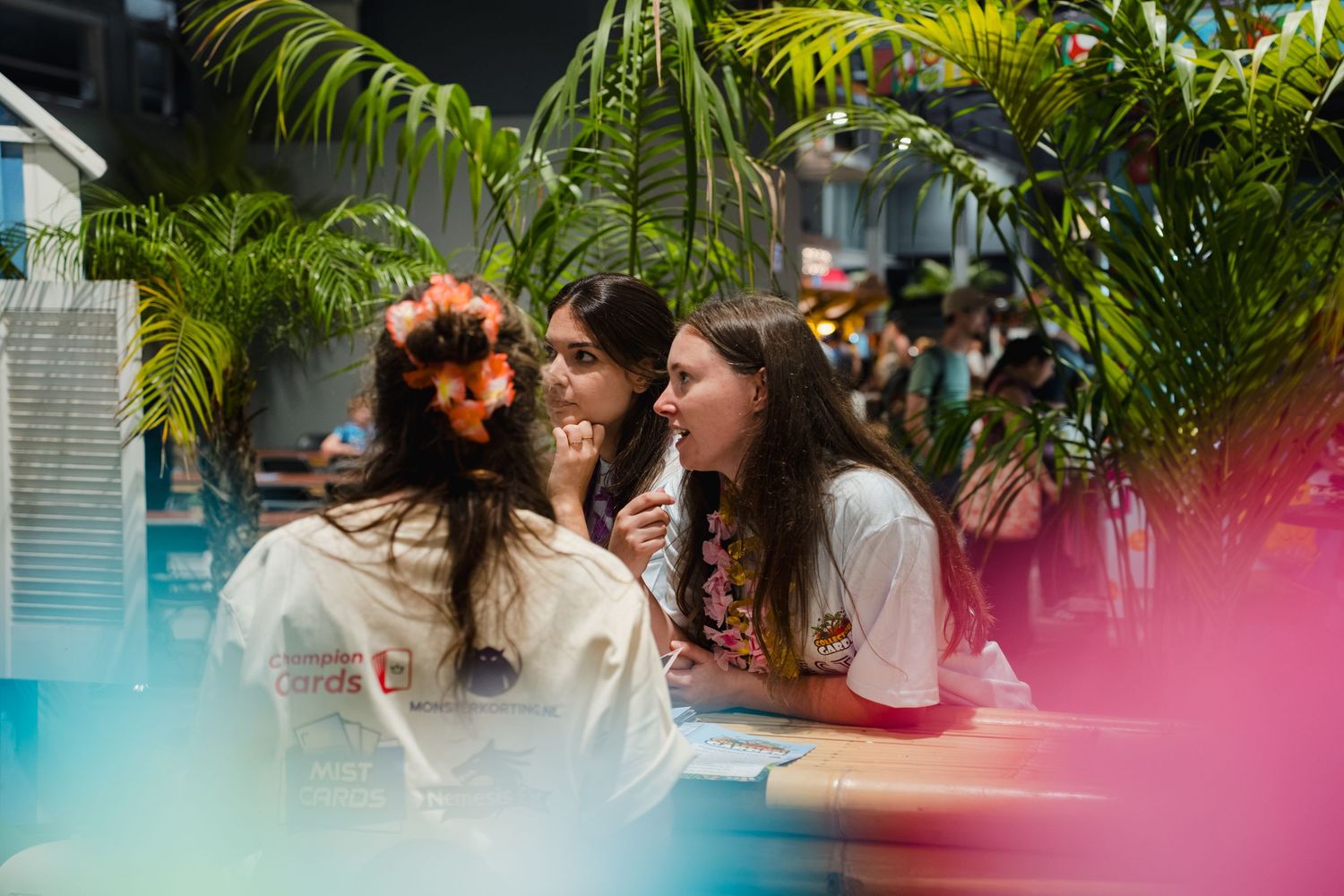 Three young women laughing and chatting together at a tropical-themed indoor event with lush green palms.