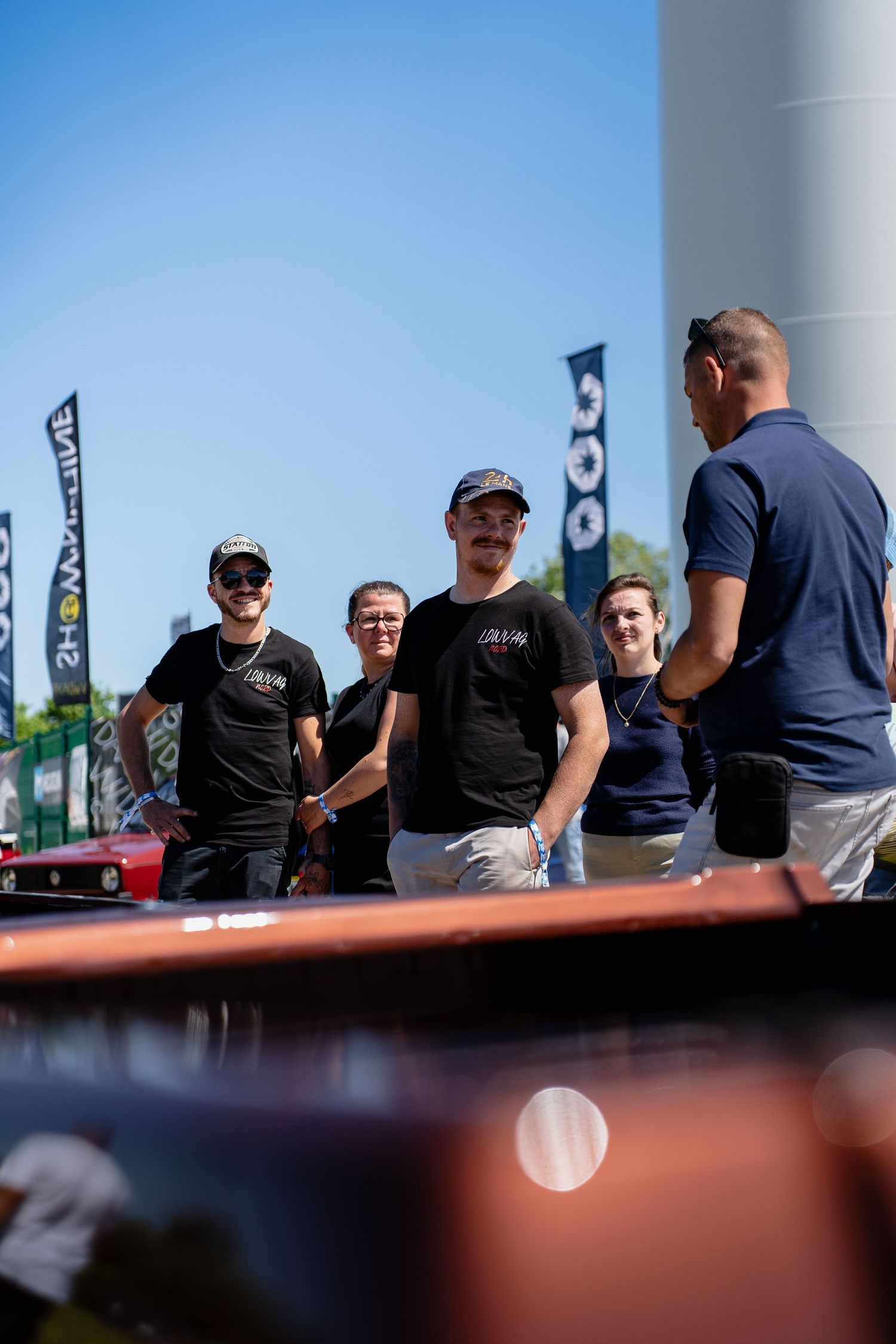 Group of people in black shirts socializing outdoors at a sunny car show event with banners in the background.