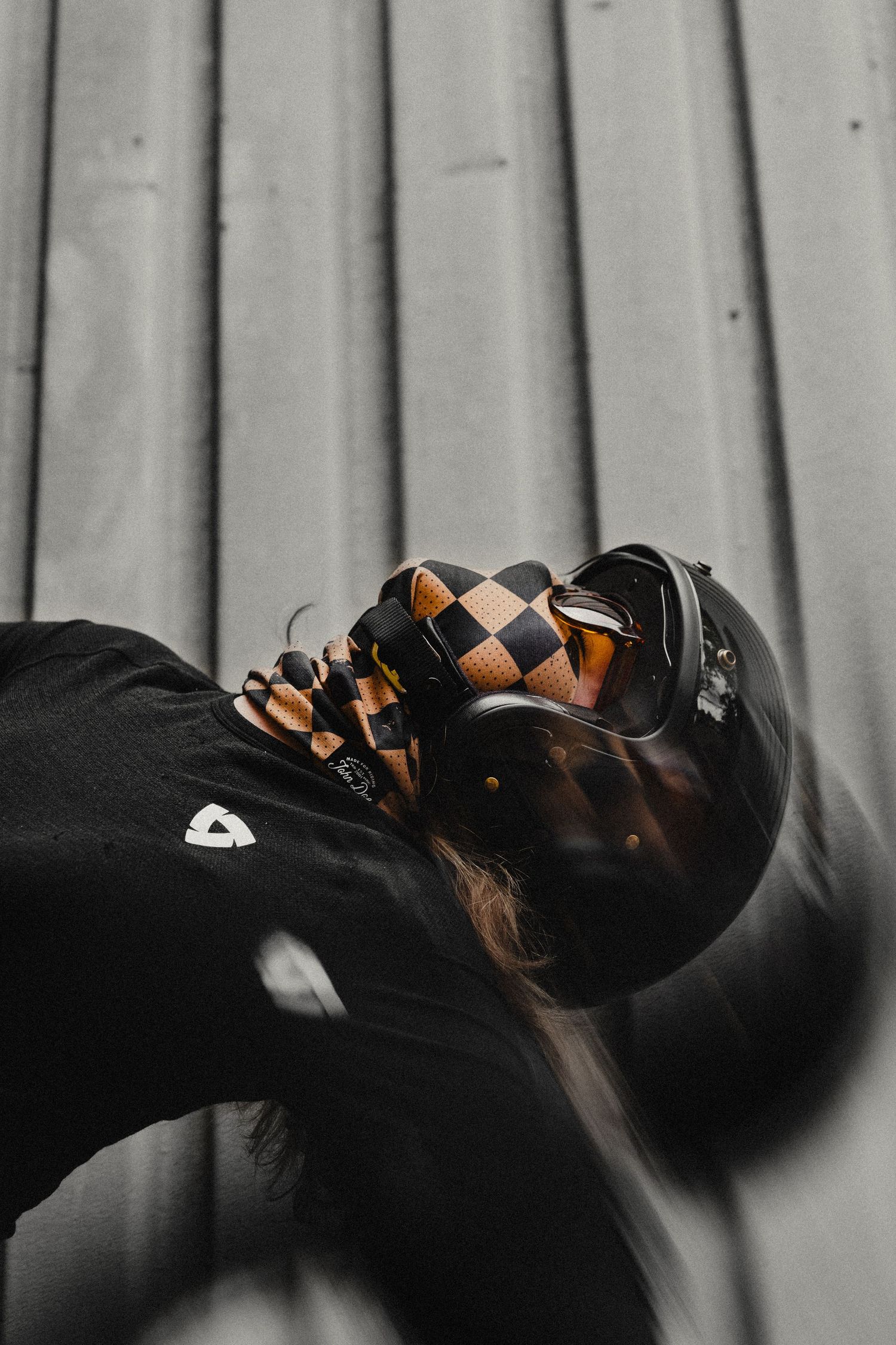 Person wearing a black t-shirt and checkered motorcycle helmet with goggles, posed against a corrugated metal wall.
