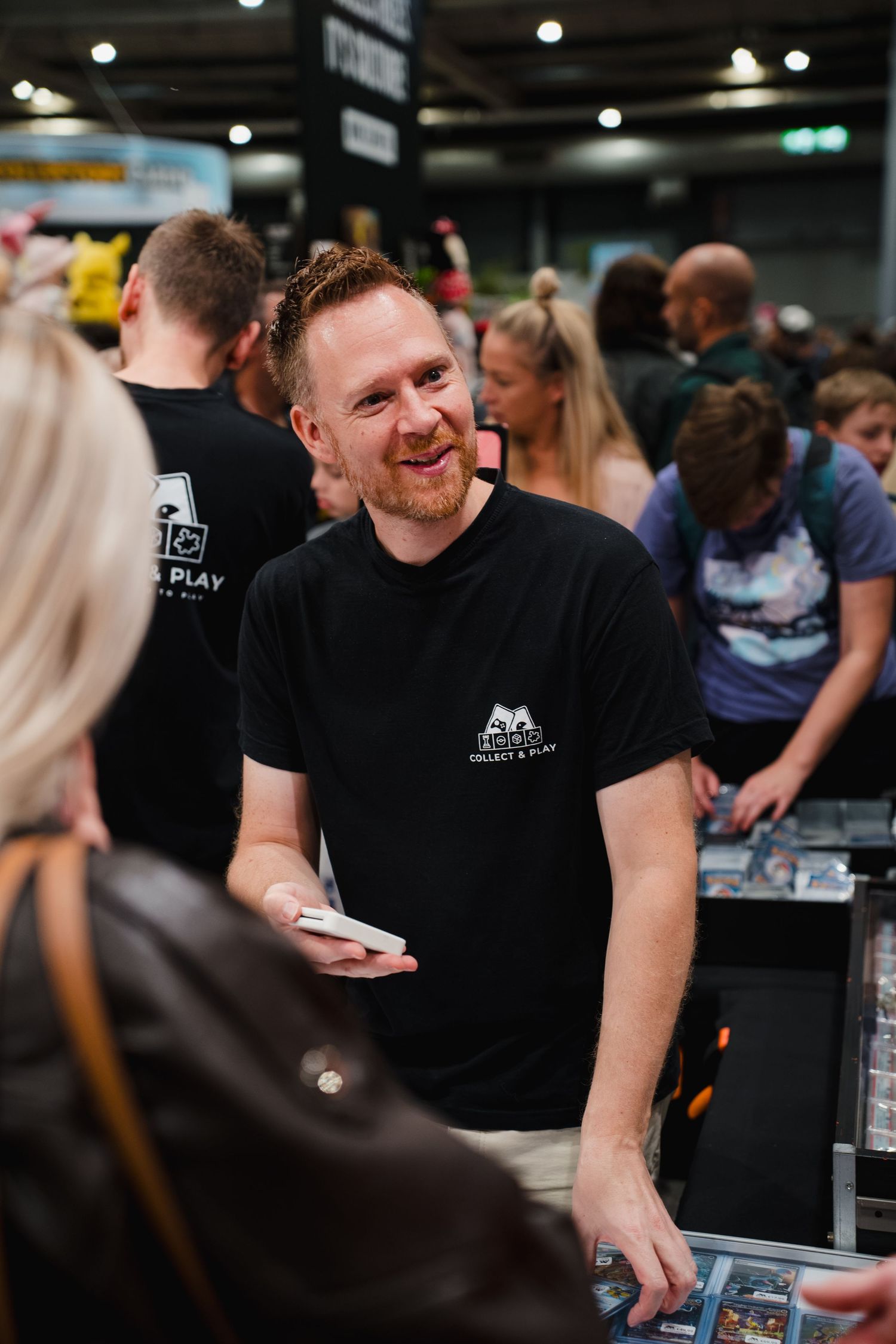 A smiling man in a black branded t-shirt holds cards at a busy gaming convention vendor booth.