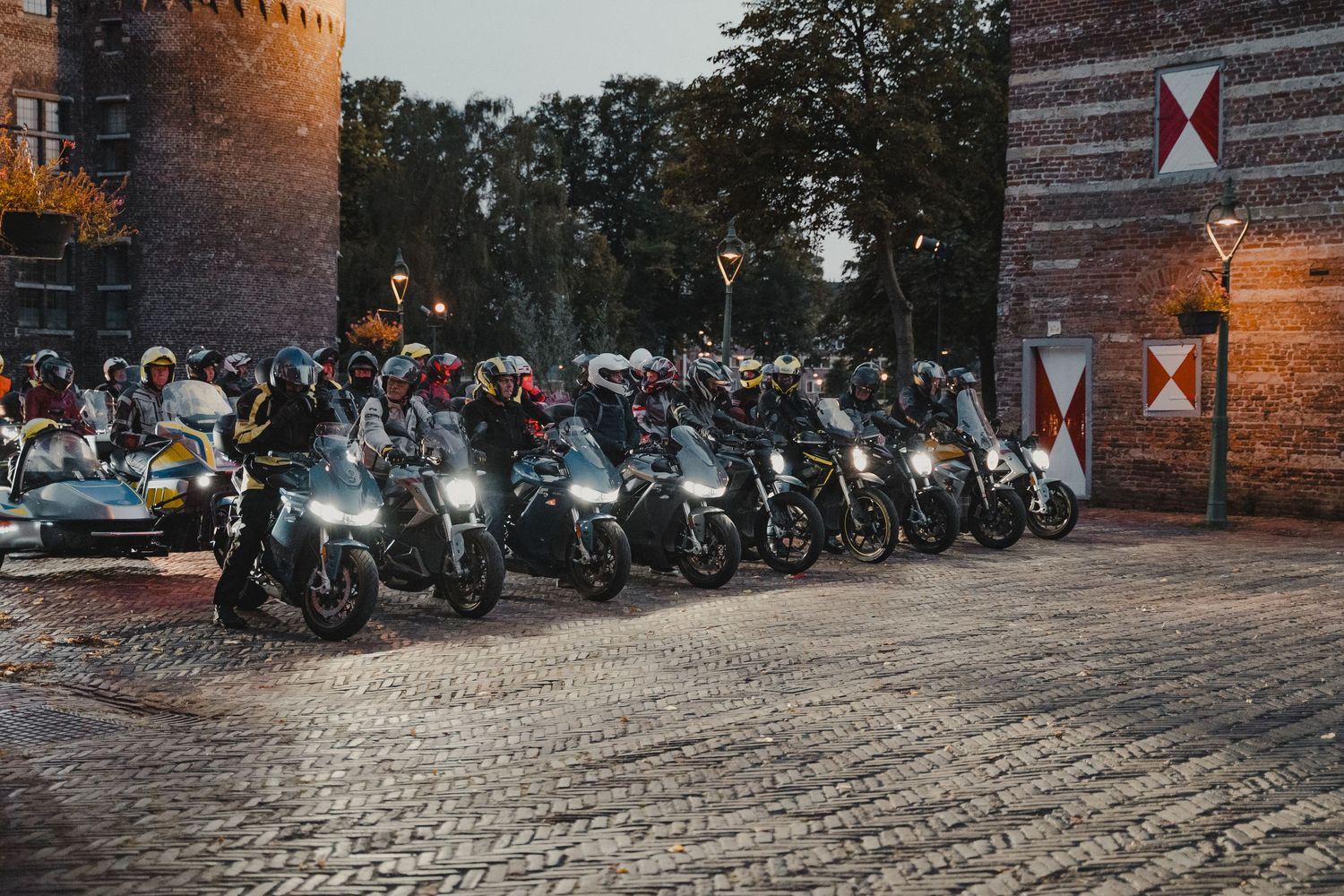 Row of parked motorcycles lined up at dusk near a historic brick tower with cobblestone ground.