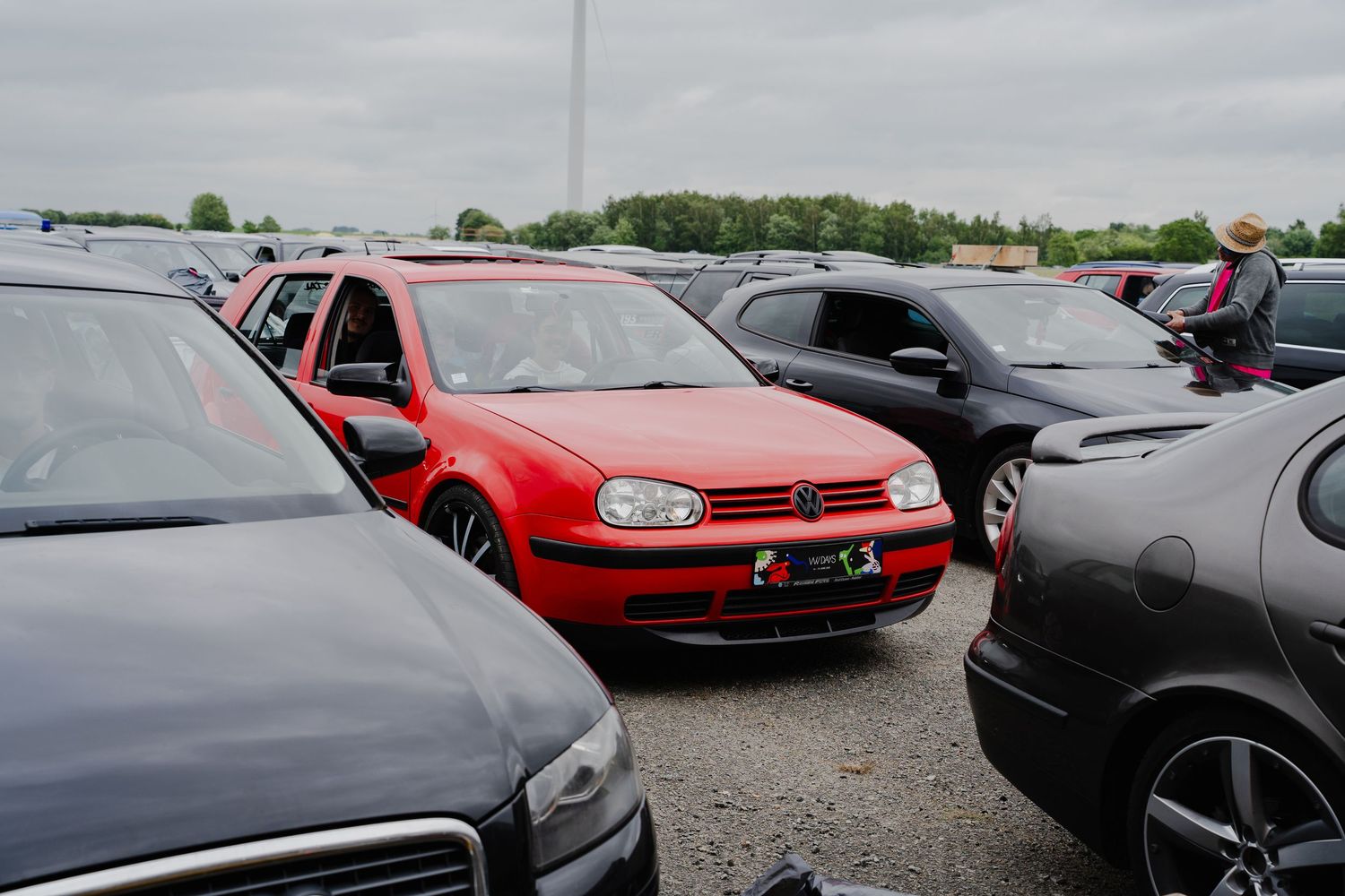 Red Volkswagen Golf parked among black cars in a crowded outdoor parking lot on an overcast day.