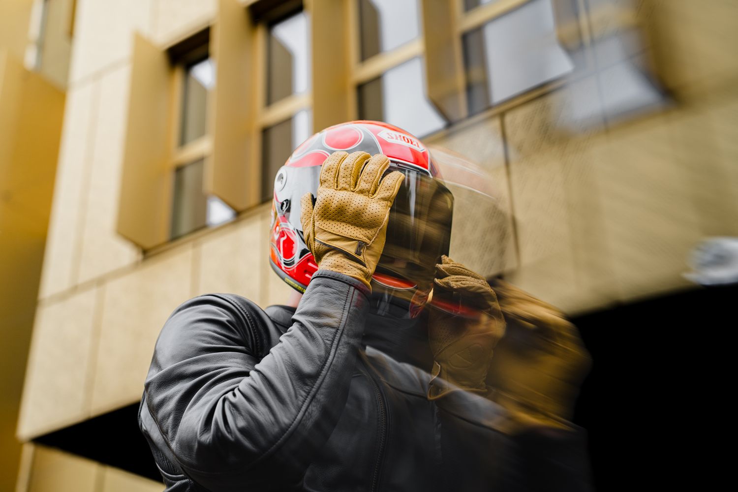 Motorcyclist putting on a red helmet with tan gloves outside a building.
