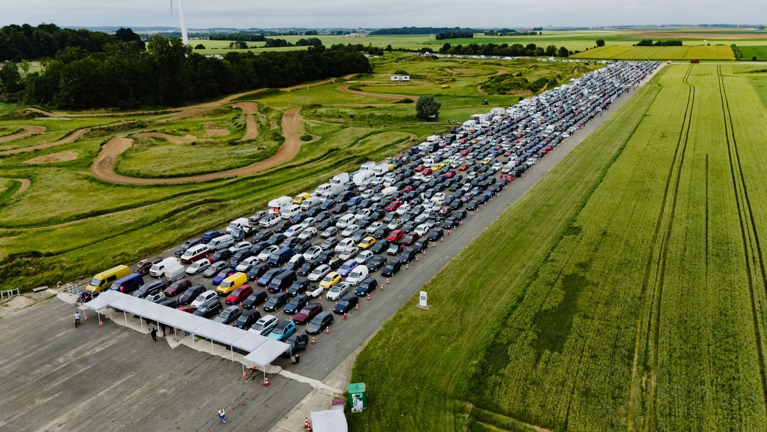 Aerial view of a large outdoor car parking lot filled with hundreds of vehicles beside green fields.