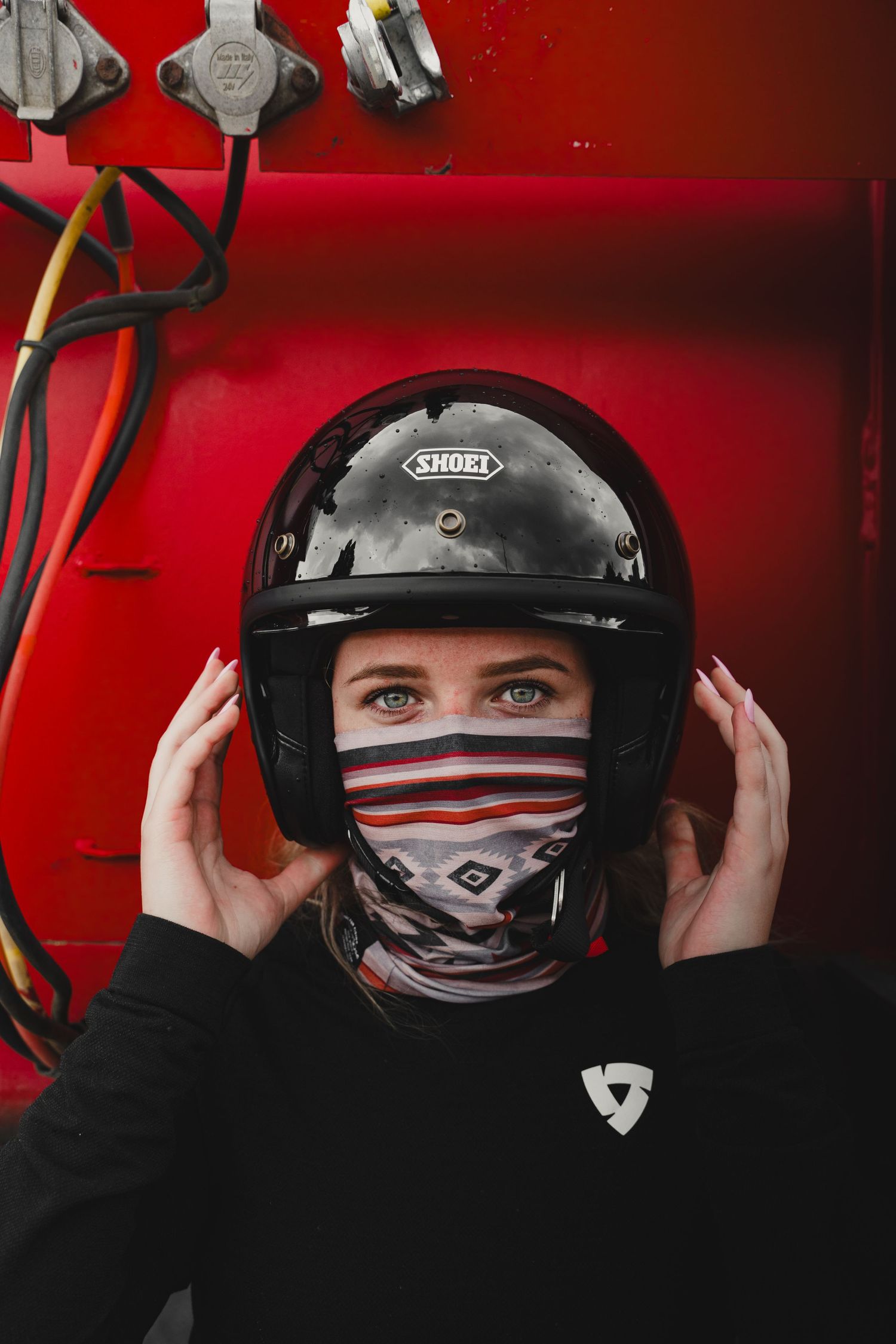 Woman wearing a black Shoei motorcycle helmet and striped face covering against a red background.