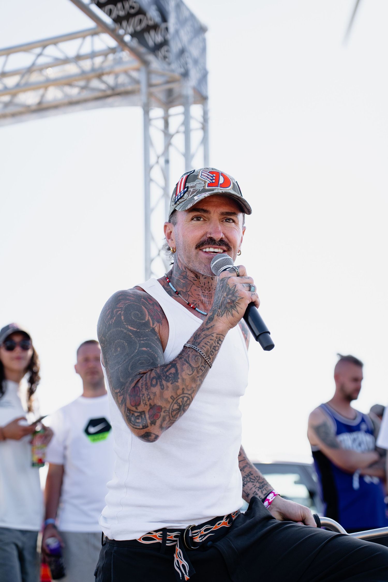 Heavily tattooed man in white tank top and baseball cap speaks into microphone at outdoor festival.