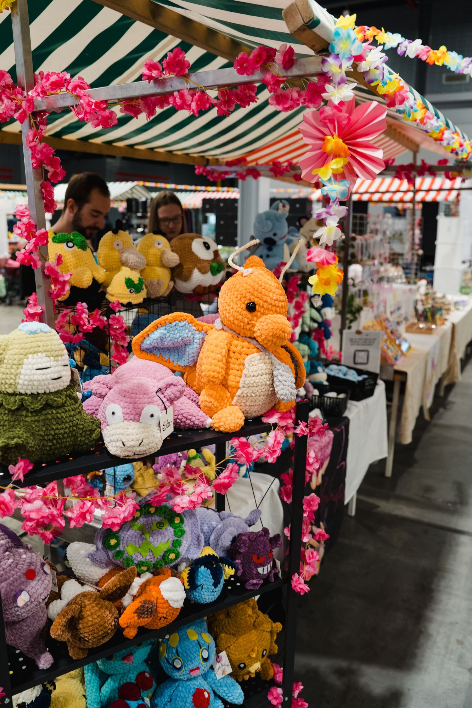 Colorful crocheted Pokemon plush toys displayed on a market stall rack decorated with pink floral garlands.
