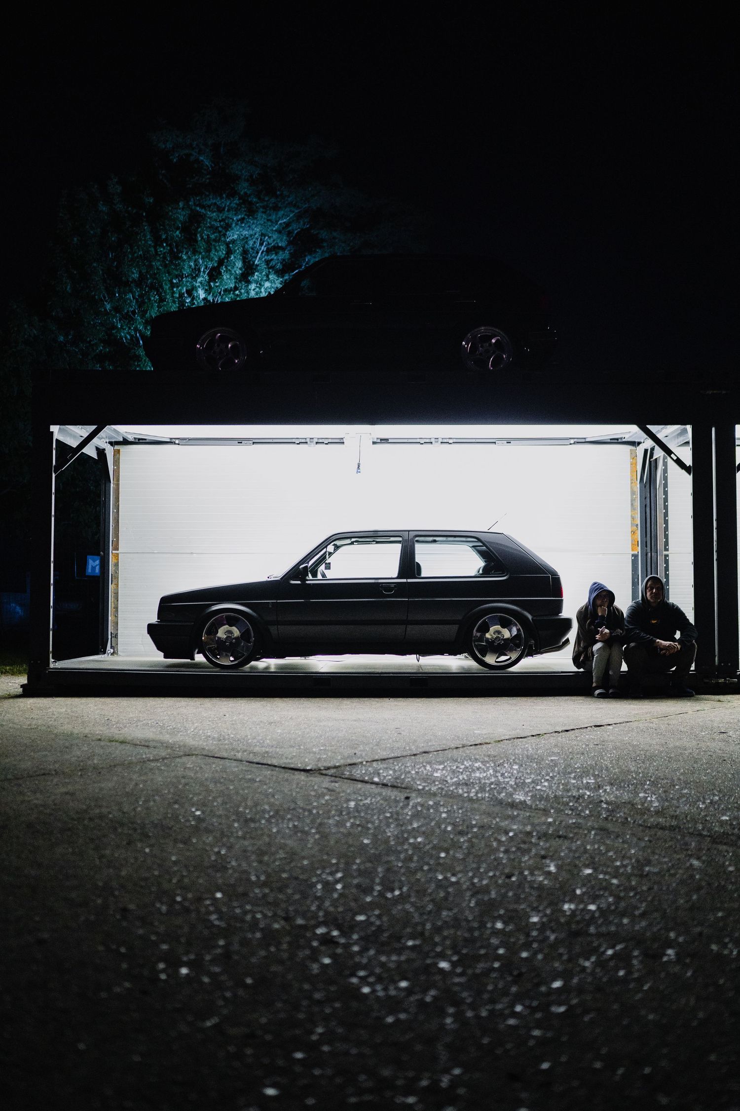 Two people crouching beside a black Volkswagen Golf in an illuminated garage at night.