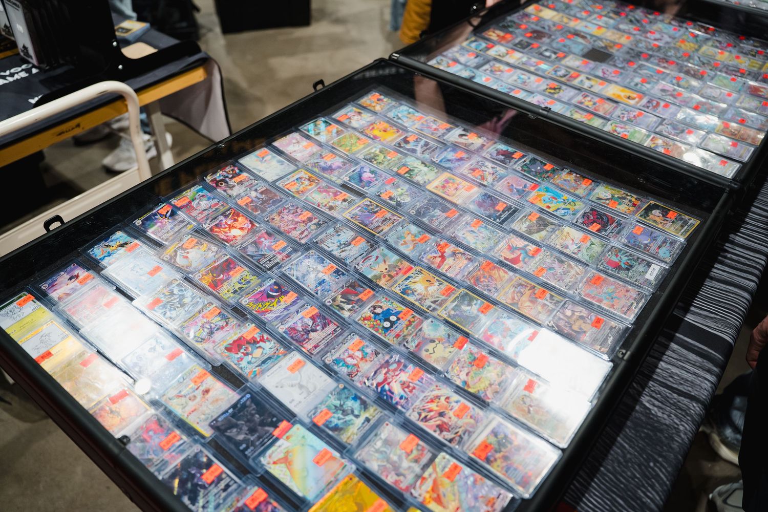 Trading card collection displayed in black cases at a convention, featuring colorful Pokemon or Yu-Gi-Oh cards in protective sleeves.