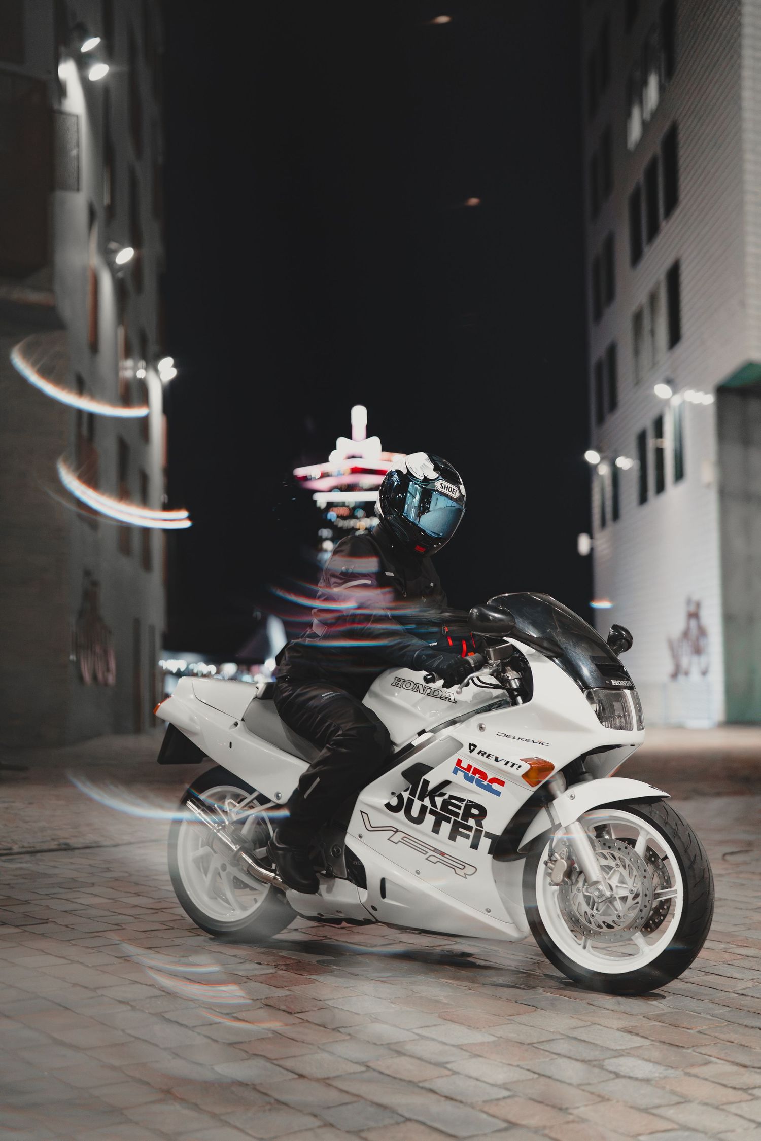 Motorcyclist on white sport bike posing at night in urban alley with light trails and city buildings.
