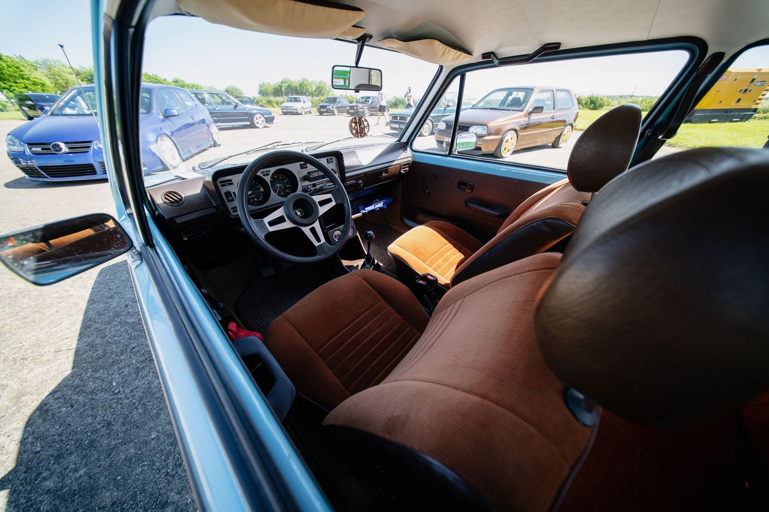 Vintage car interior with brown leather seats and classic dashboard, viewed from open door in sunny parking lot.