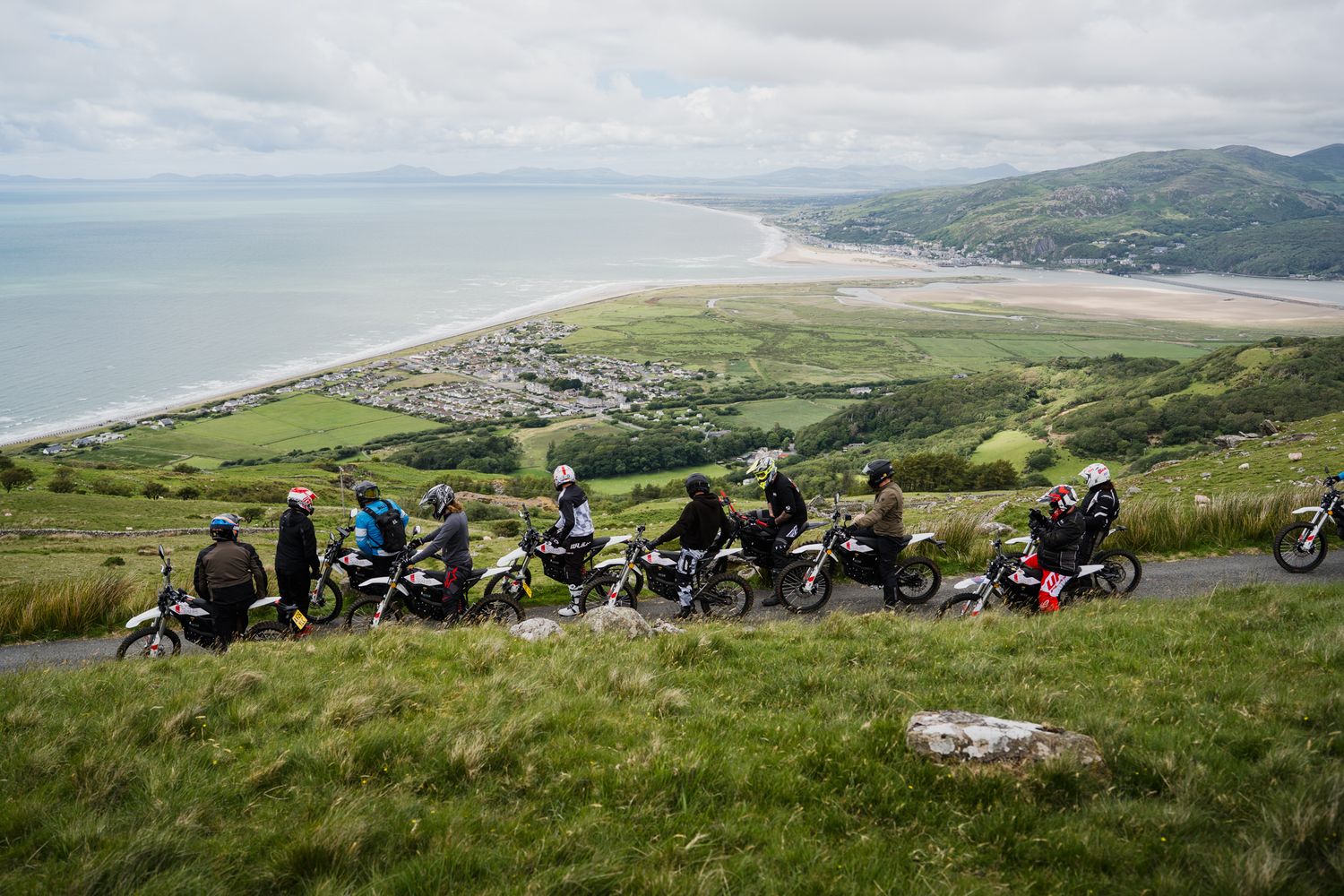 Group of motorcyclists parked on a hillside overlooking a scenic coastal beach and green valley landscape.