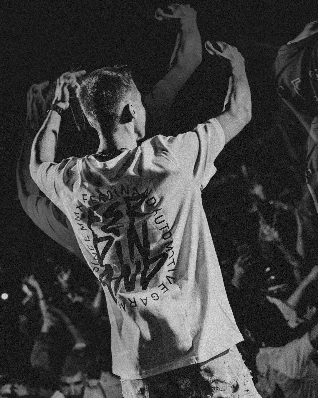 Concert performers seen from behind wearing white shirts with text during a live show.