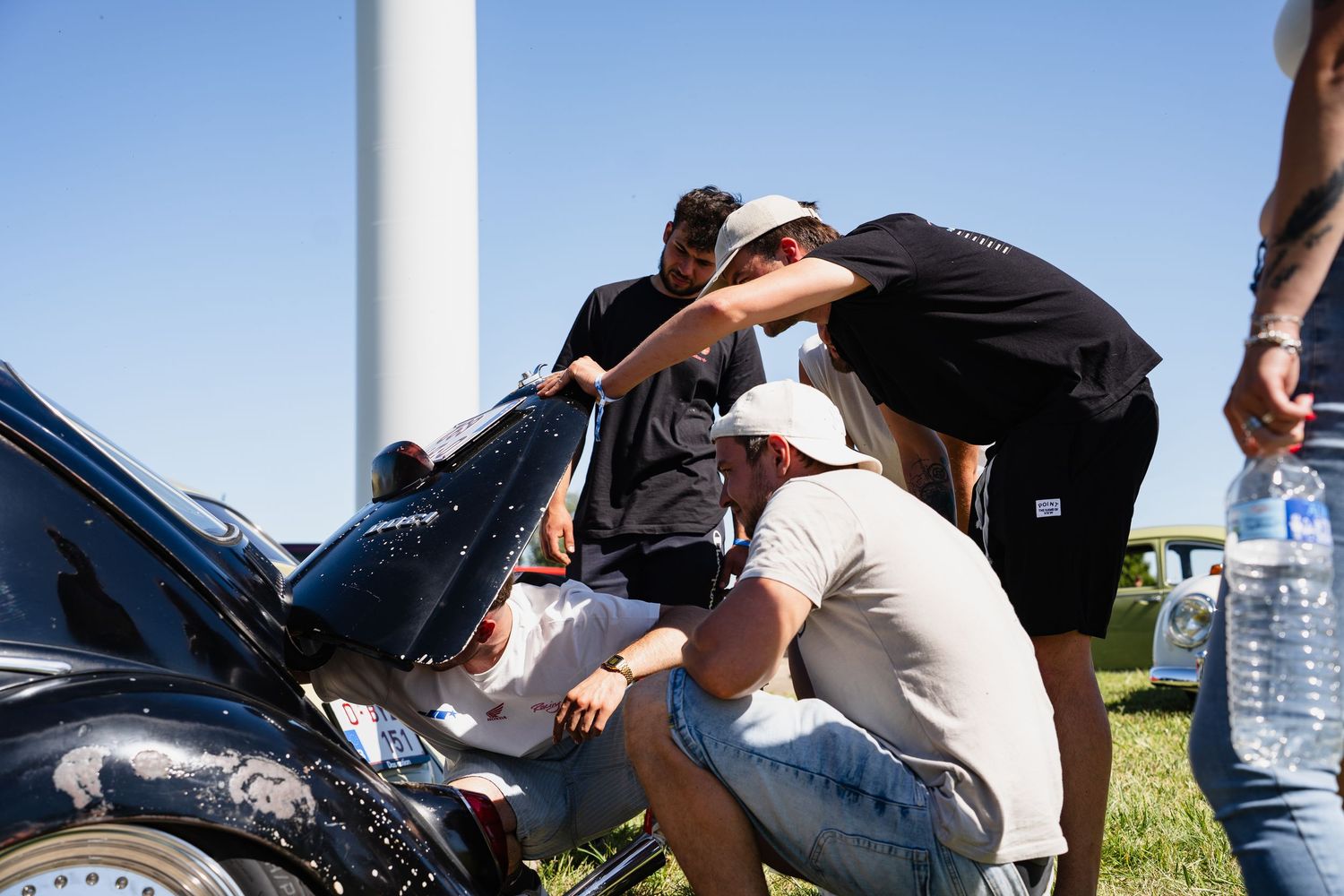 Group of men examining a black car engine at an outdoor car show near a wind turbine on a sunny day.