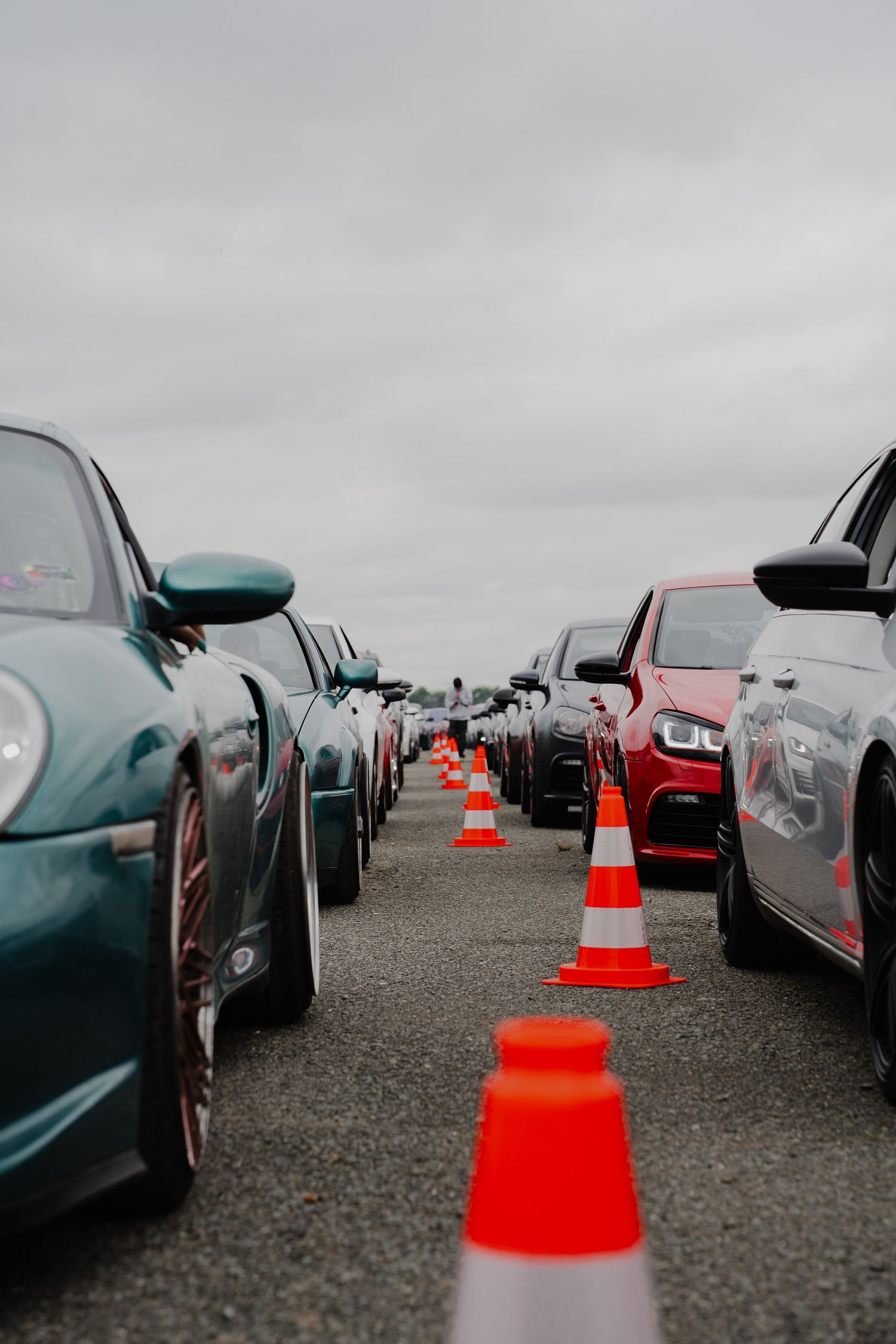 Rows of cars lined up with orange traffic cones between them on a cloudy day at an automotive event.