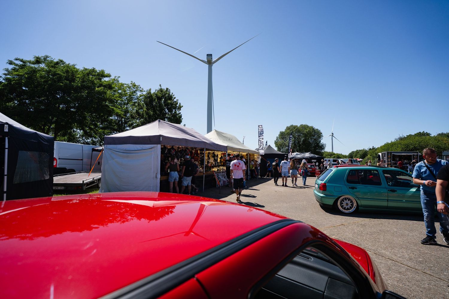 Outdoor car meet event with white tents, wind turbine, and colorful vehicles parked under a clear blue sky.