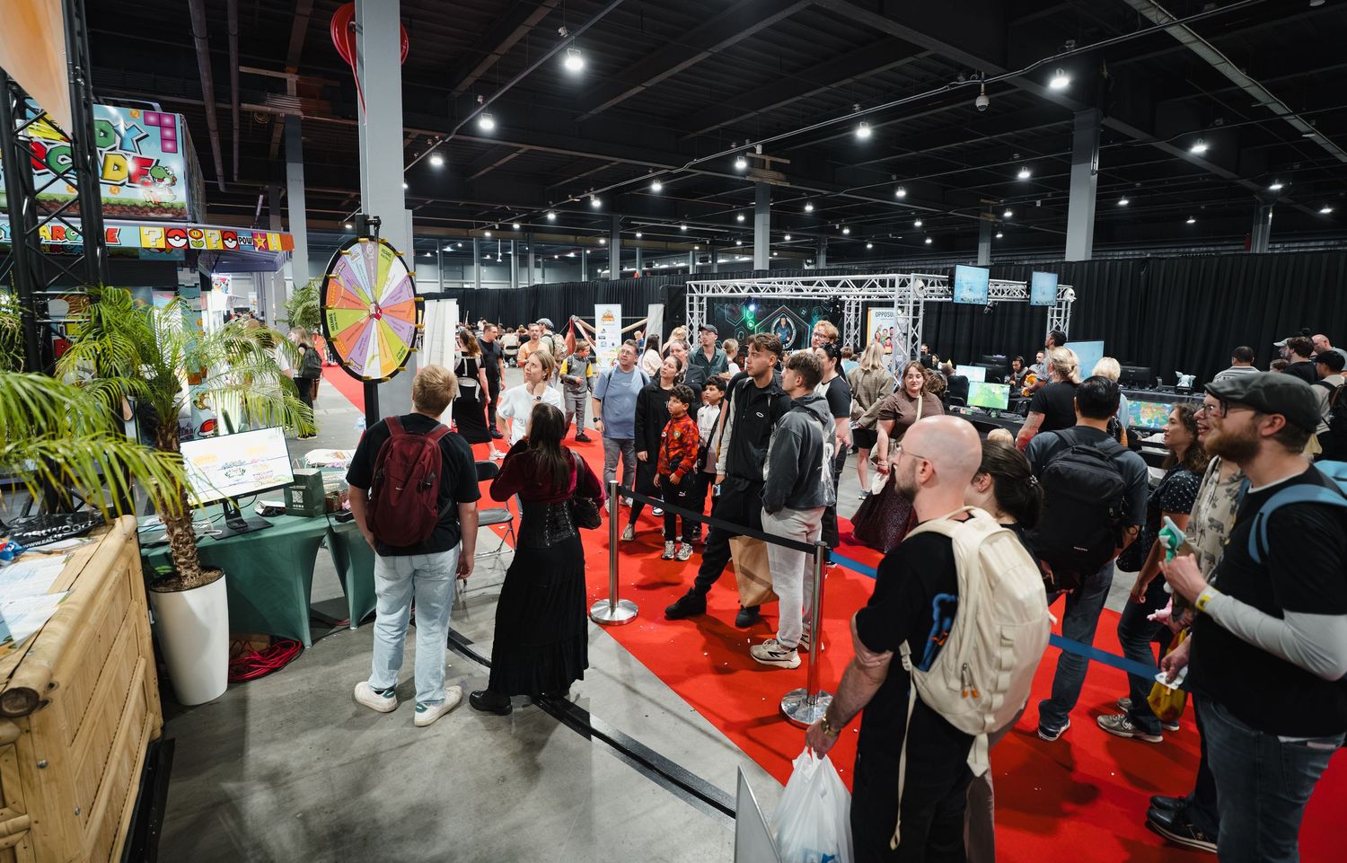 Crowded convention hall with attendees browsing vendor booths, a prize wheel, and displays under bright overhead lights.