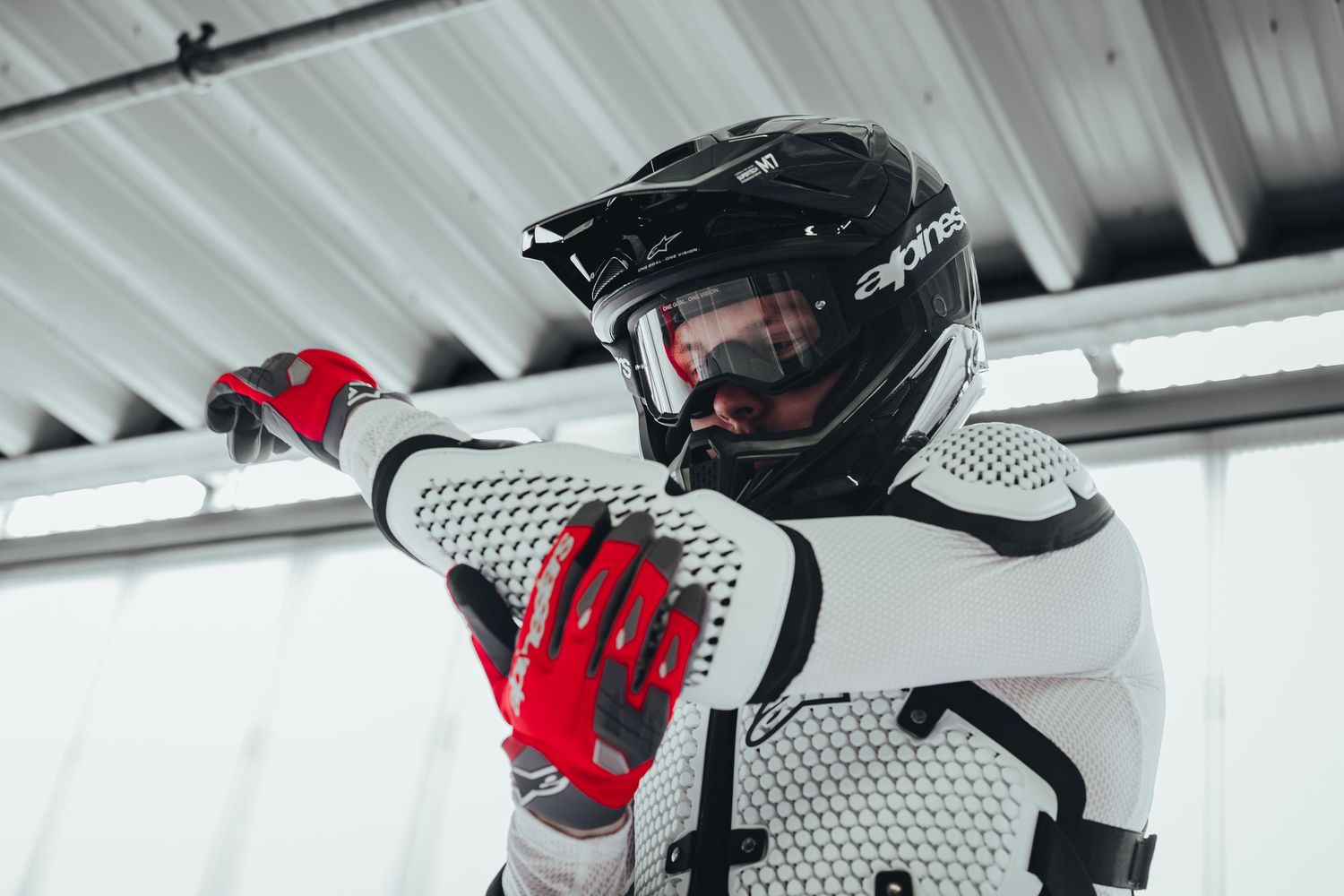 Motocross rider in white Alpinestars gear, red gloves, and black helmet with goggles adjusting equipment indoors.