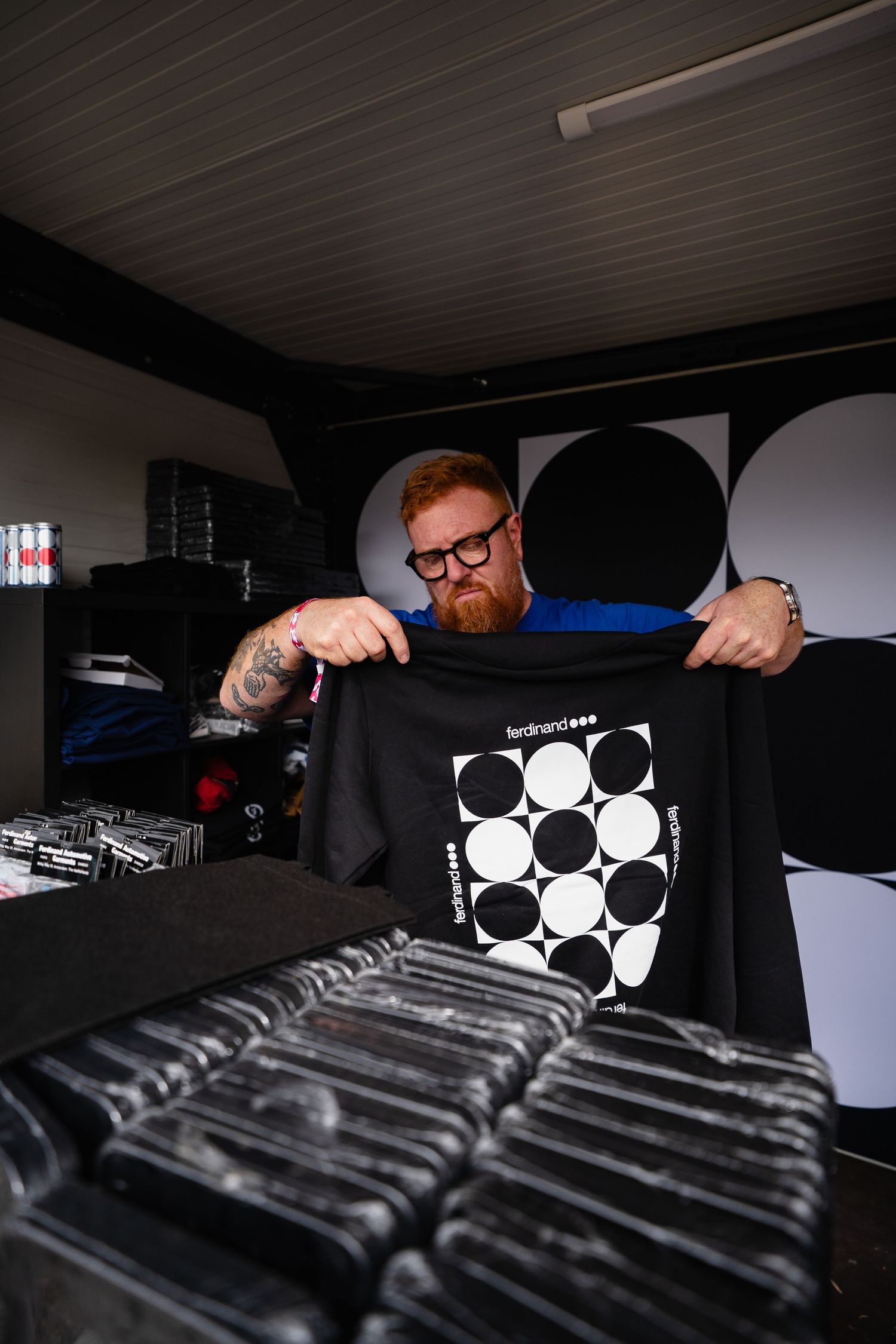 Bearded man with glasses holding a black Ferdinand t-shirt with bold circle graphic design at a merchandise stand.