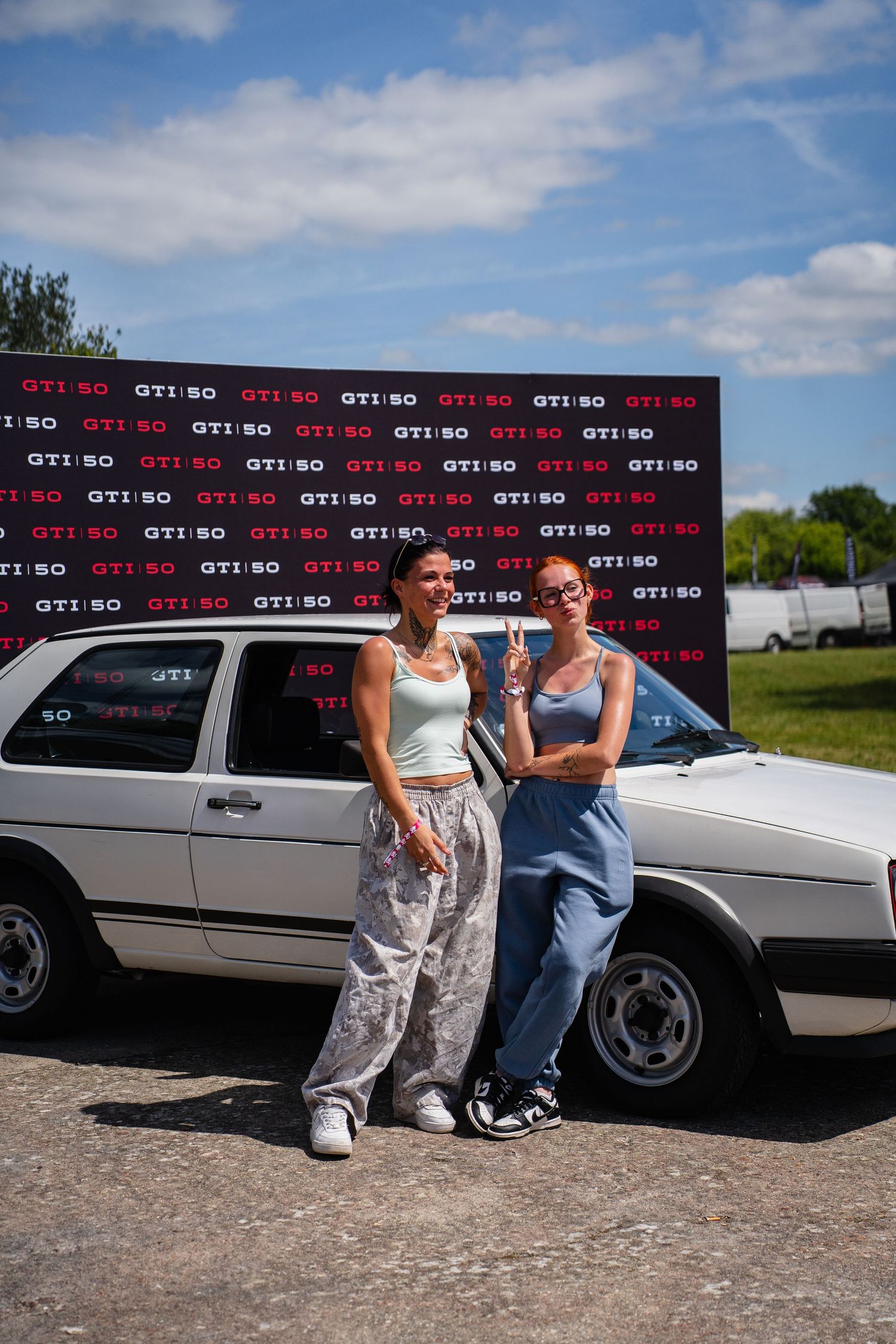 Two women posing in front of a white VW GTI car at a GTI 50th anniversary event on a sunny day.