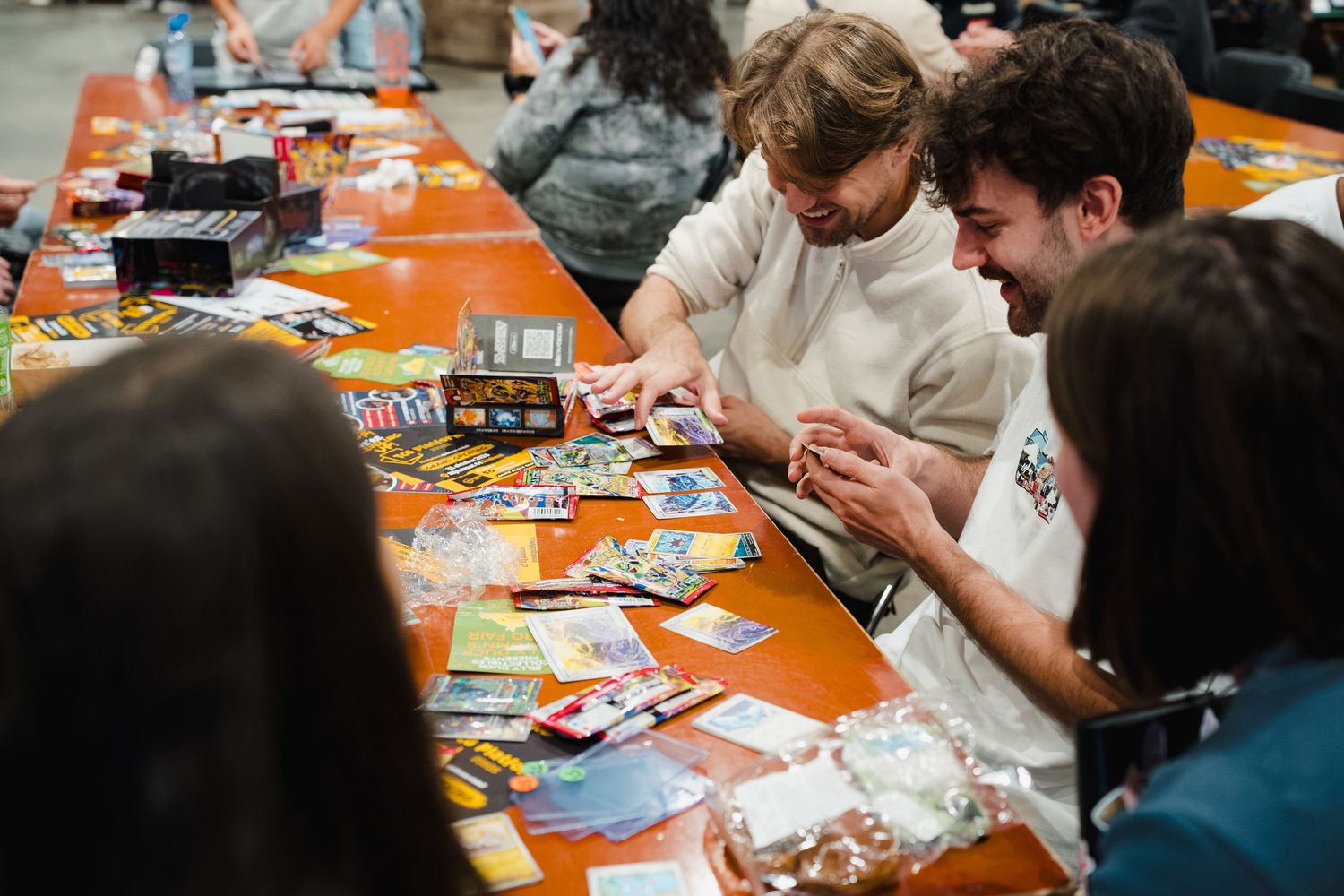 People playing card games at an orange table covered with colorful cards and game pieces at a gaming event.