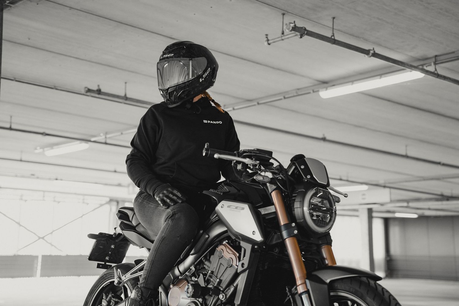 A motorcyclist in a black helmet and jacket sits on a dark cruiser motorcycle in a concrete parking garage.
