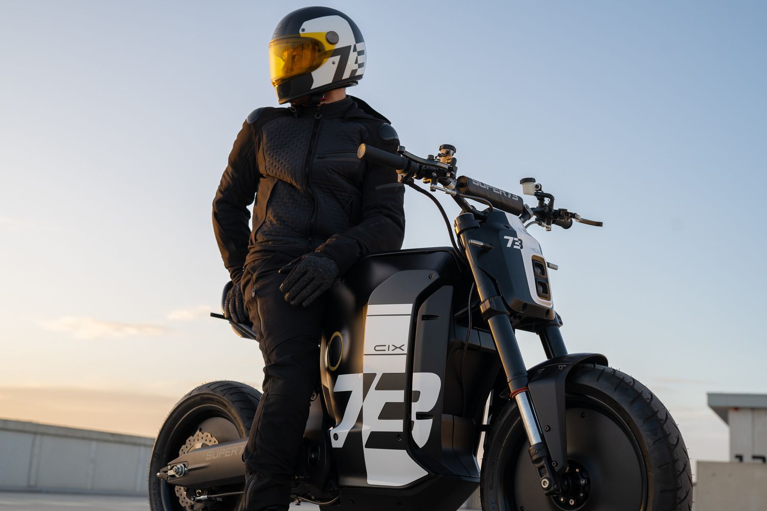 Motorcycle rider in black gear standing next to modern electric bike at sunset.